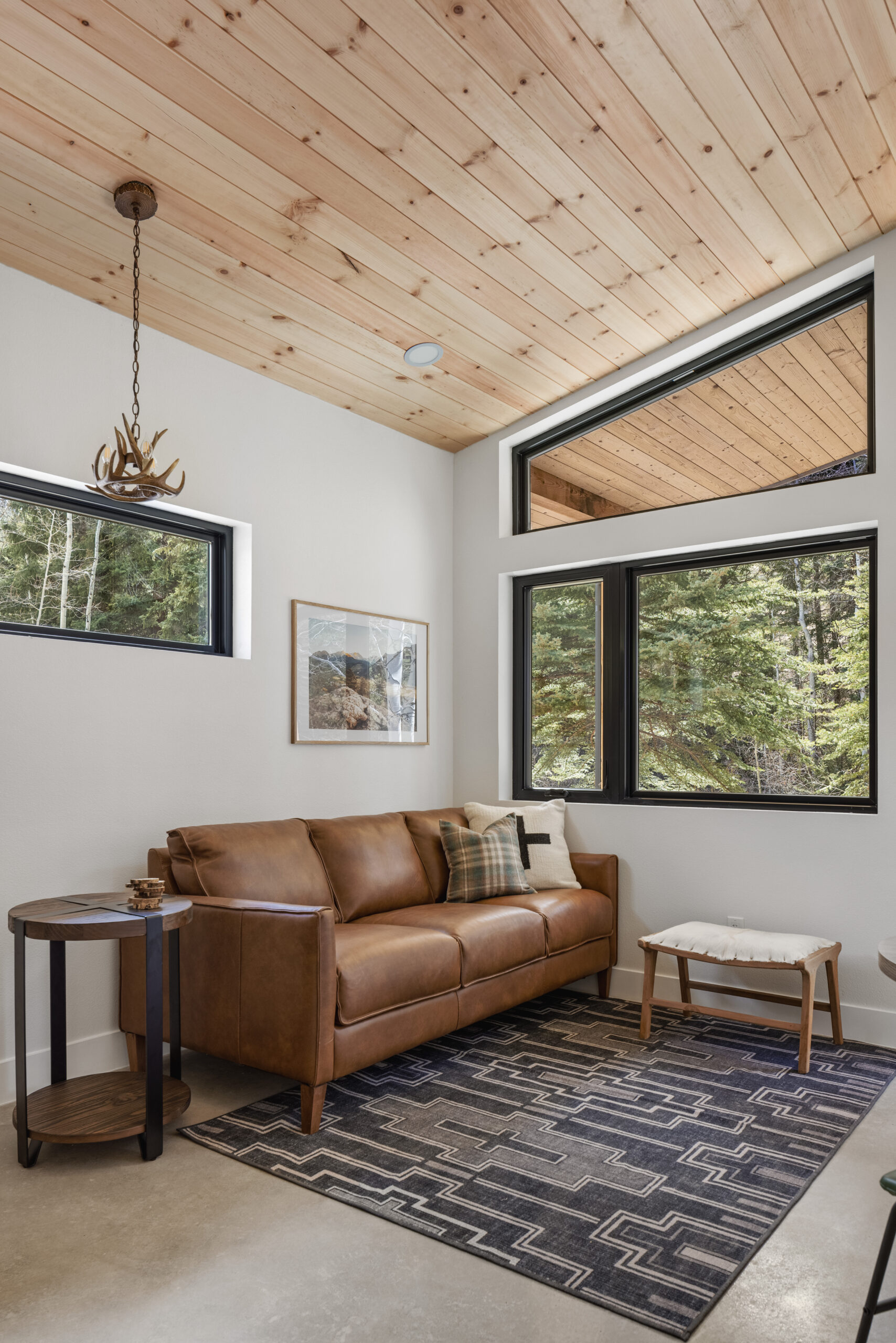 A cozy living room with a brown leather sofa, patterned rug, round wooden side table, and ottoman. Large windows show trees outside. The space has light walls and a wood-paneled ceiling with an antler-style light fixture.