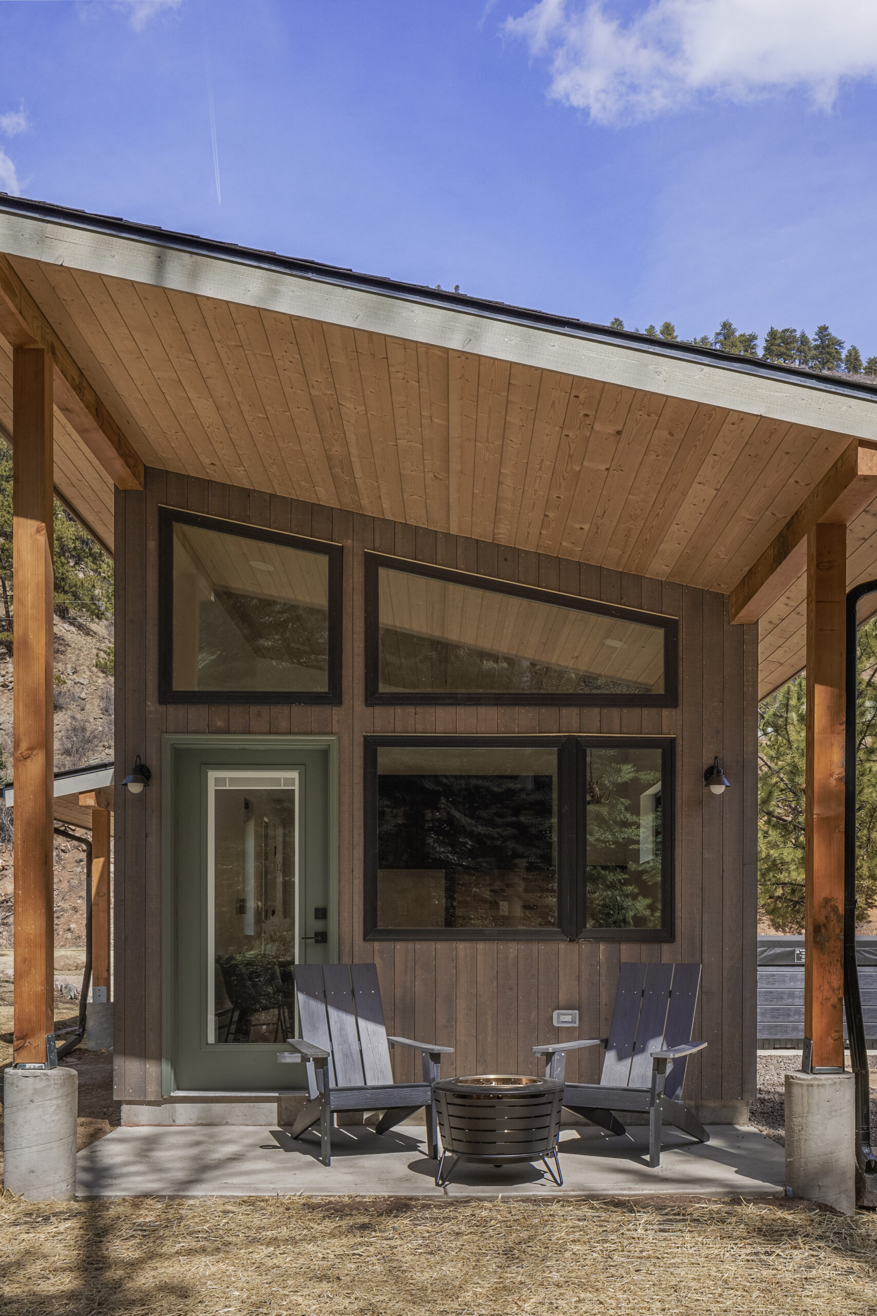A modern cabin with large windows and a slanted wooden roof features two Adirondack chairs and a fire pit on the front patio. Trees and blue sky are visible in the background.