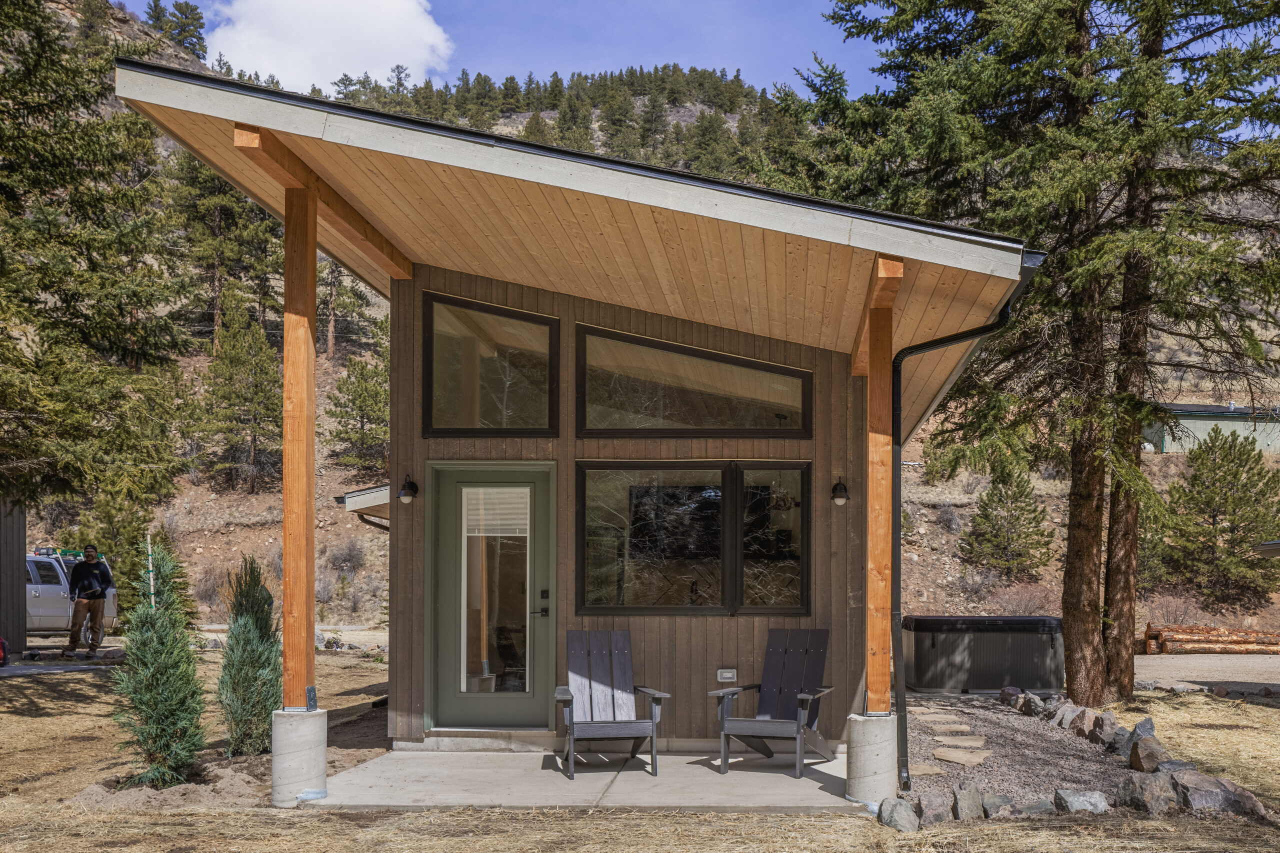 A modern small cabin with a sloped roof and large windows, two wooden chairs on a concrete porch, surrounded by pine trees and mountains in the background.