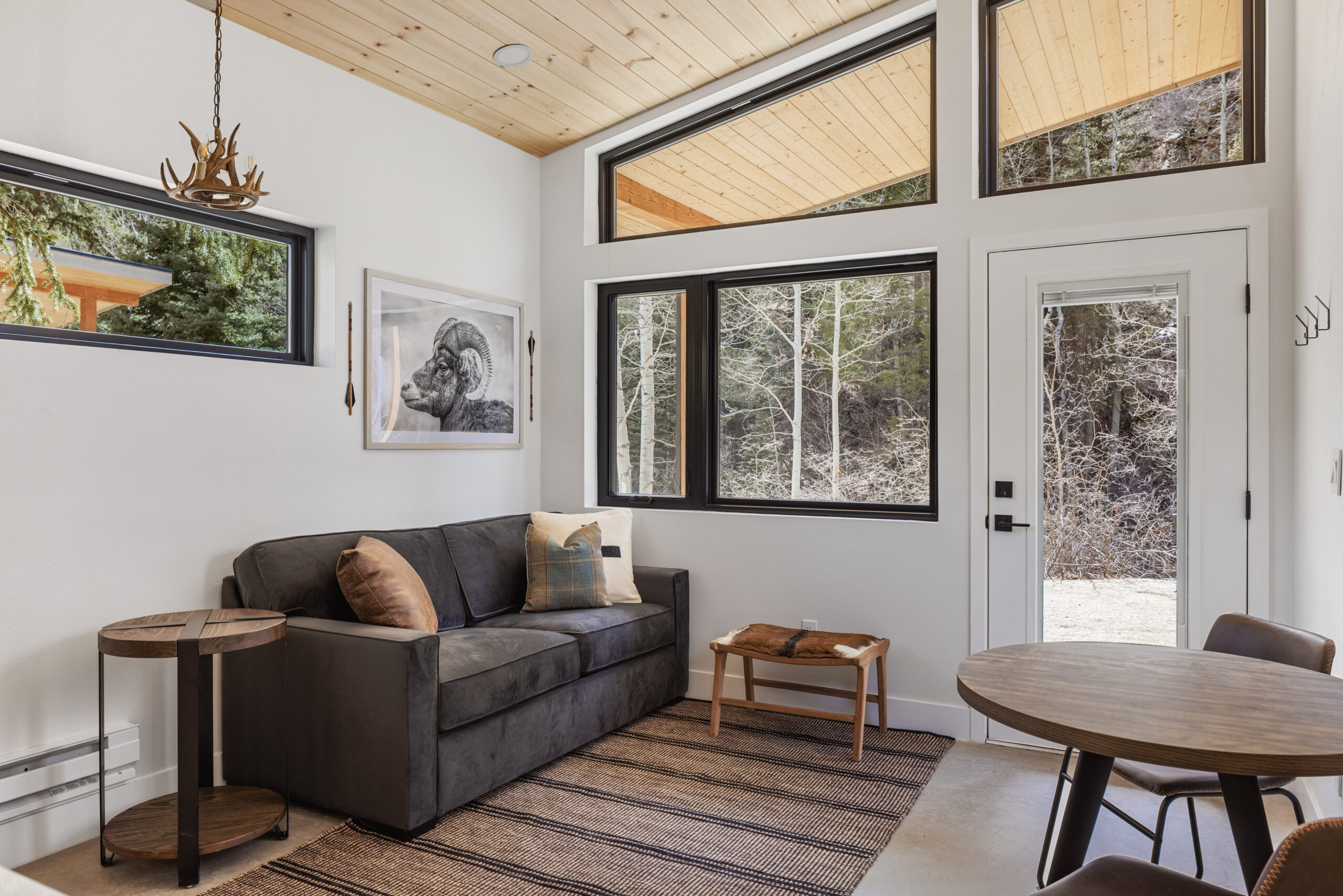 Modern, cozy living room with a gray sofa, rustic wood tables, a round dining table with chairs, large windows showing trees outside, and decor like a framed animal print and an antler-style light fixture.