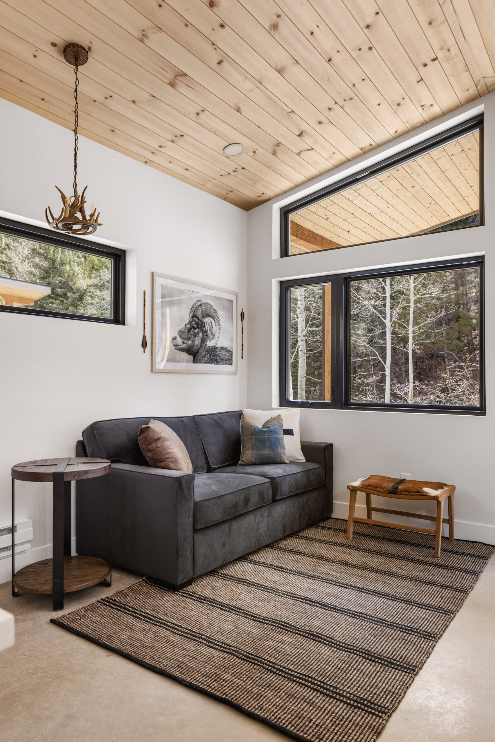 A cozy modern sitting area with a dark gray sofa, striped rug, small wooden table, antler-style pendant light, and large windows showing a snowy forest. A framed animal artwork hangs on the wall.