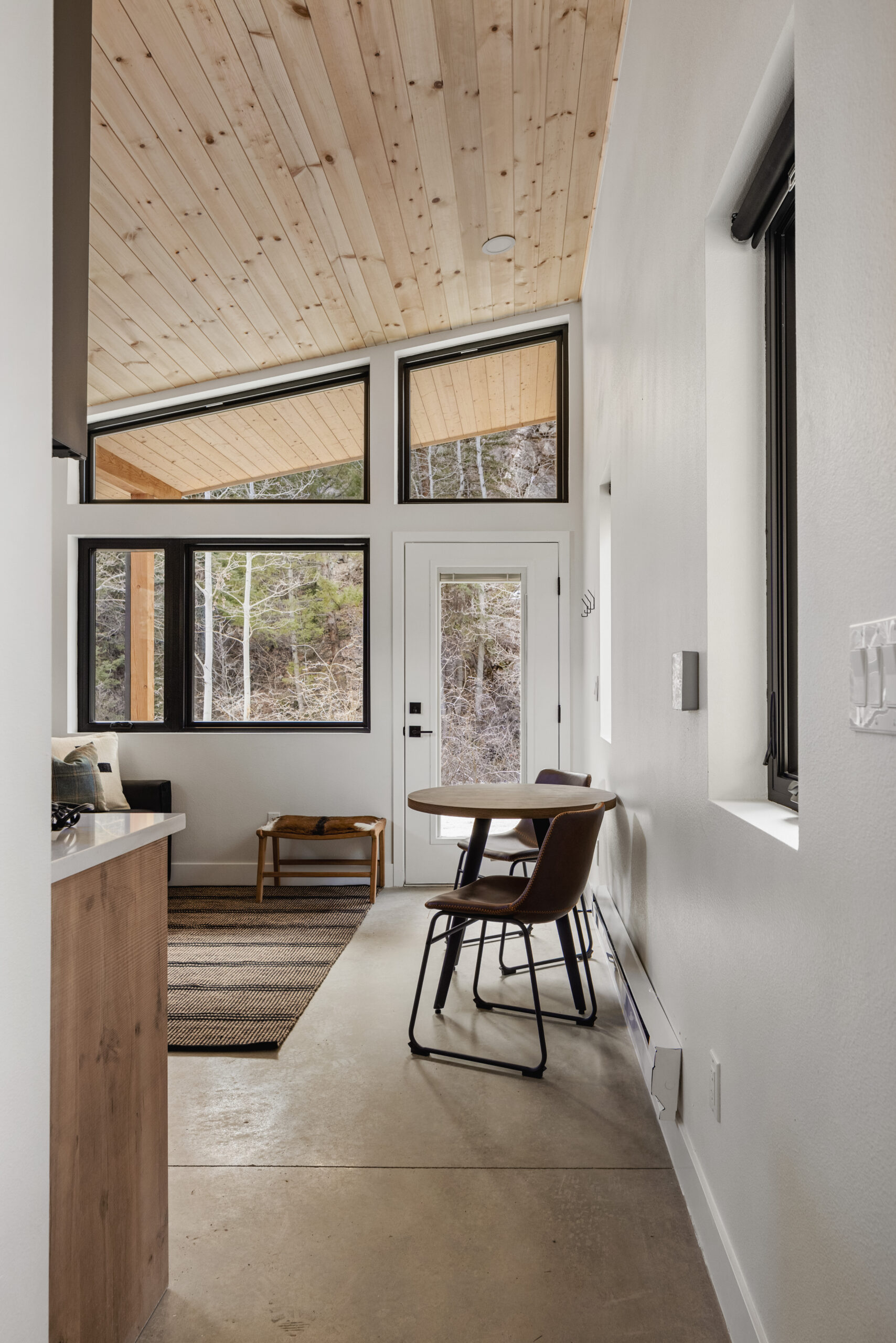Small, modern room with a light wood ceiling, white walls, and large windows. A round table with two chairs sits near the door, while a bench and rug are by the window. Trees are visible outside.