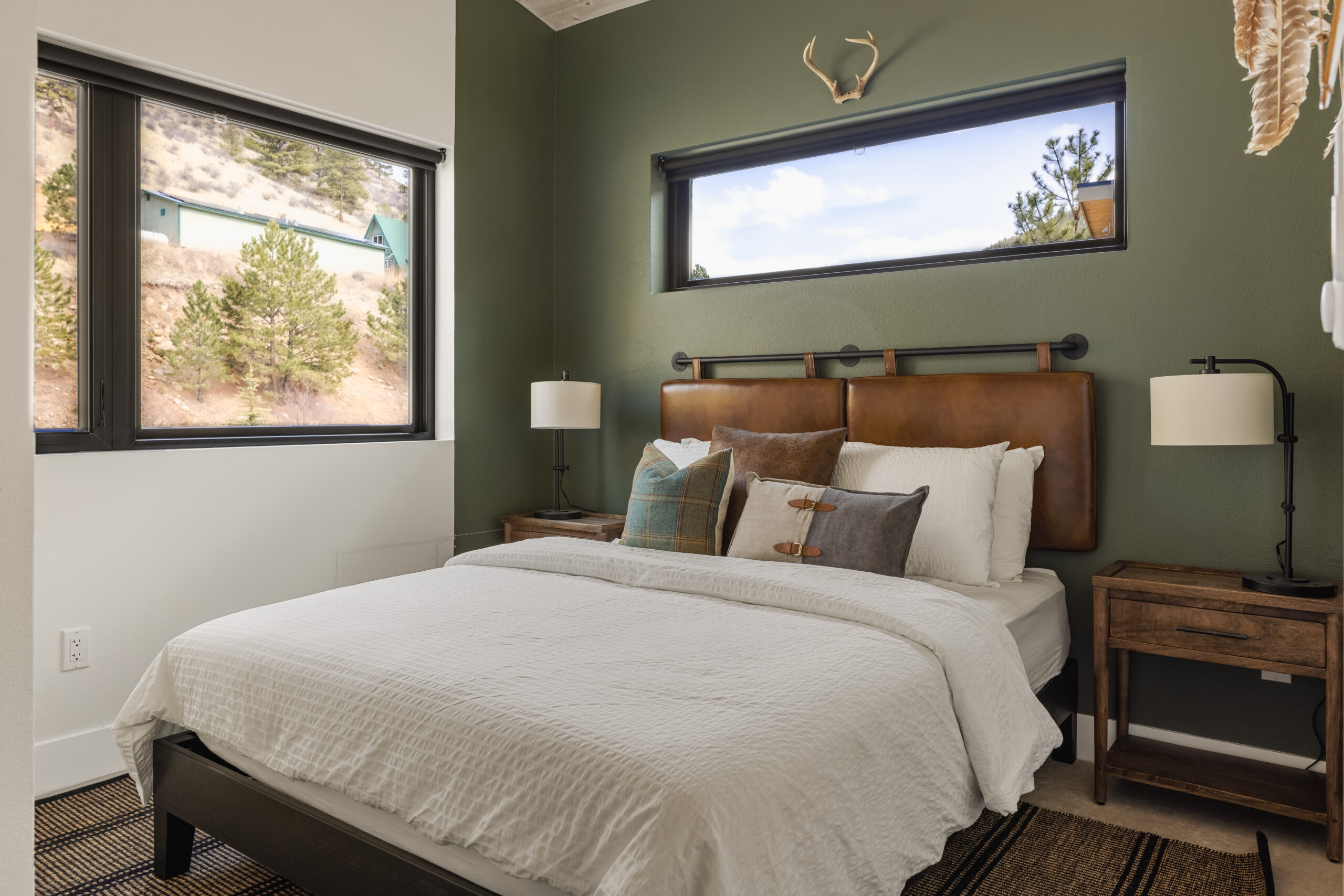 Cozy bedroom with green accent wall, brown leather headboard, white bedding, and assorted pillows. Two wooden nightstands with lamps, windows with outdoor views, and antlers mounted above the bed.