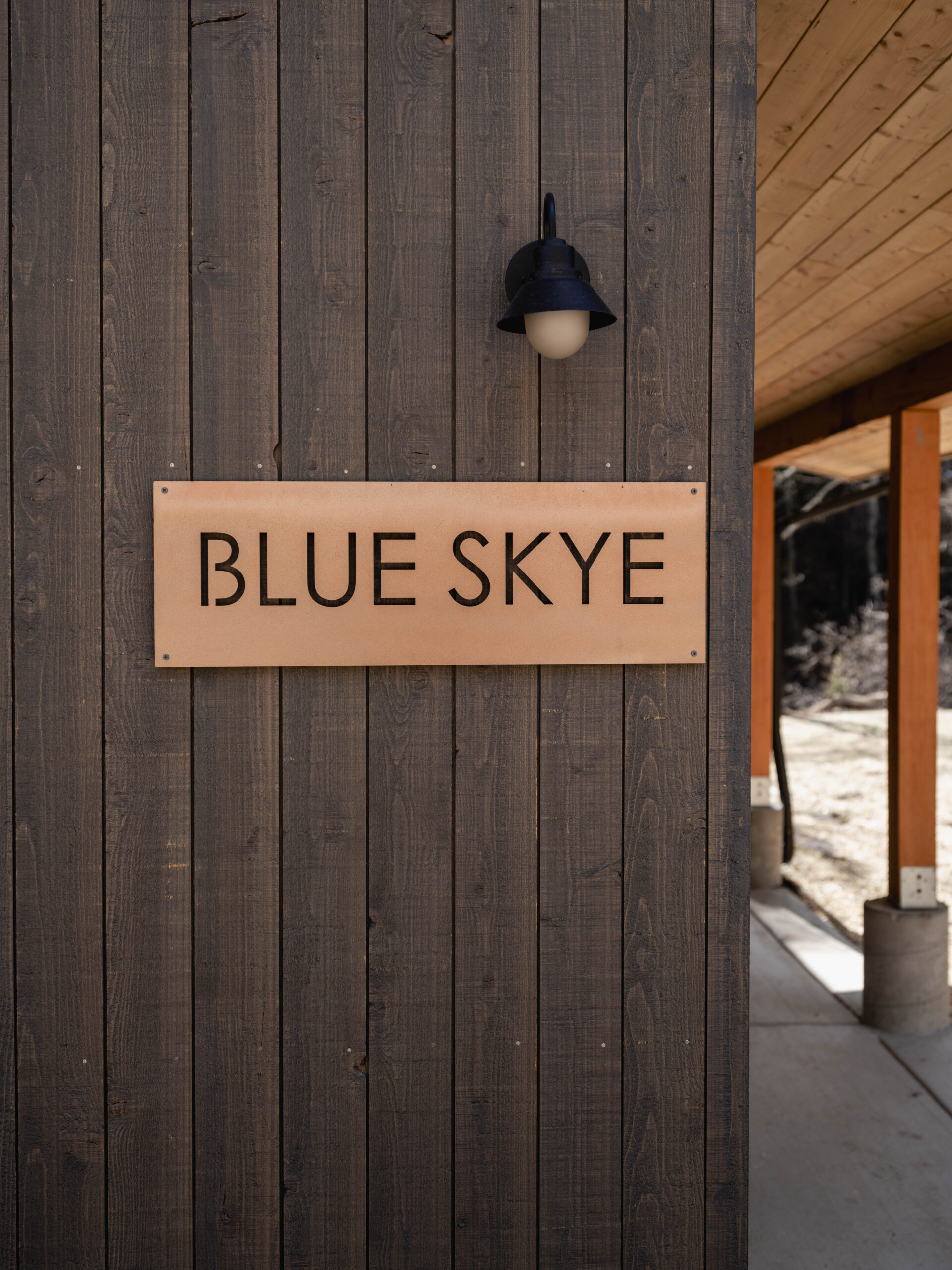 A wooden wall with a metal sign reading “BLUE SKYE” below a black outdoor light fixture; part of a porch with wooden beams is visible to the right.