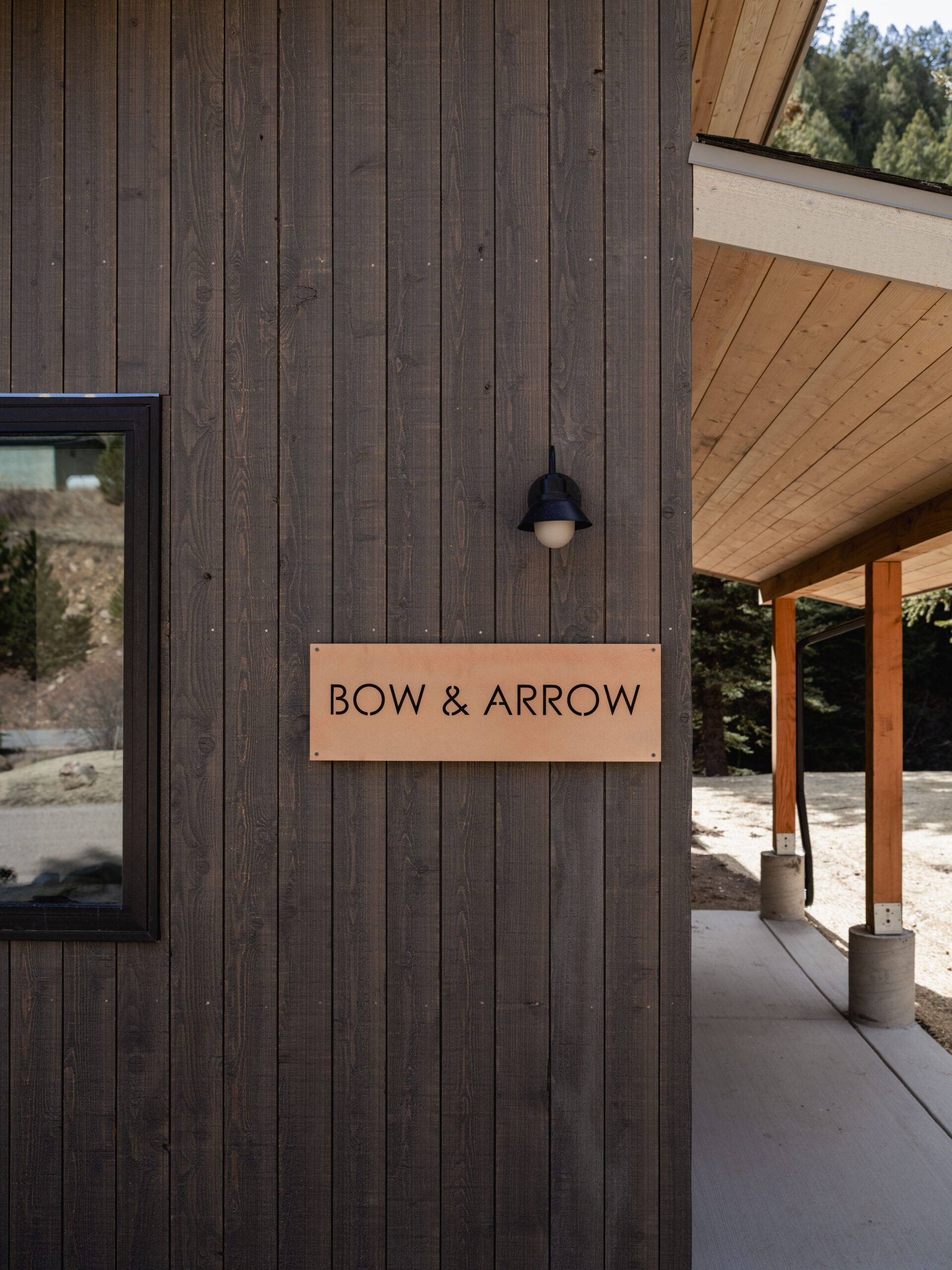 A wooden building exterior features a sign that reads "BOW & ARROW" mounted on dark vertical siding, with a window, outdoor light, and a covered walkway supported by wooden posts. Trees are visible in the background.