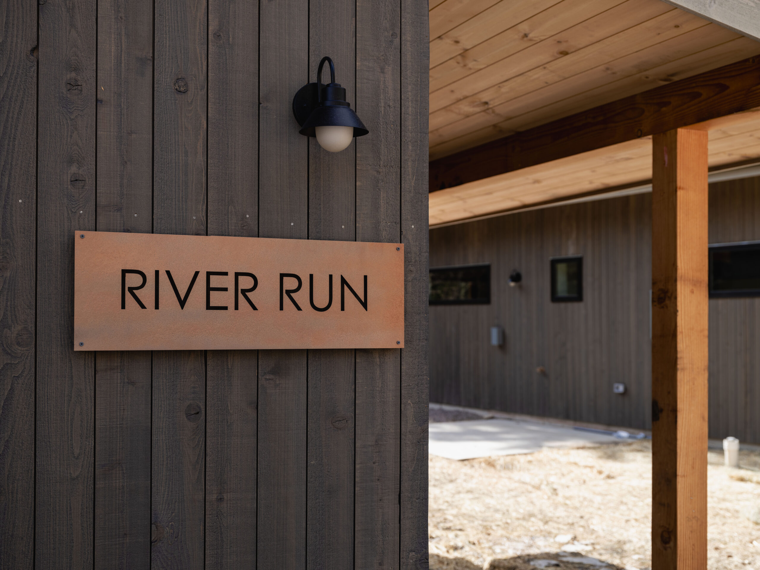 A wooden building exterior features a metal sign that reads "RIVER RUN." A black outdoor light fixture is mounted above the sign, and a covered walkway with wooden posts is visible on the right.