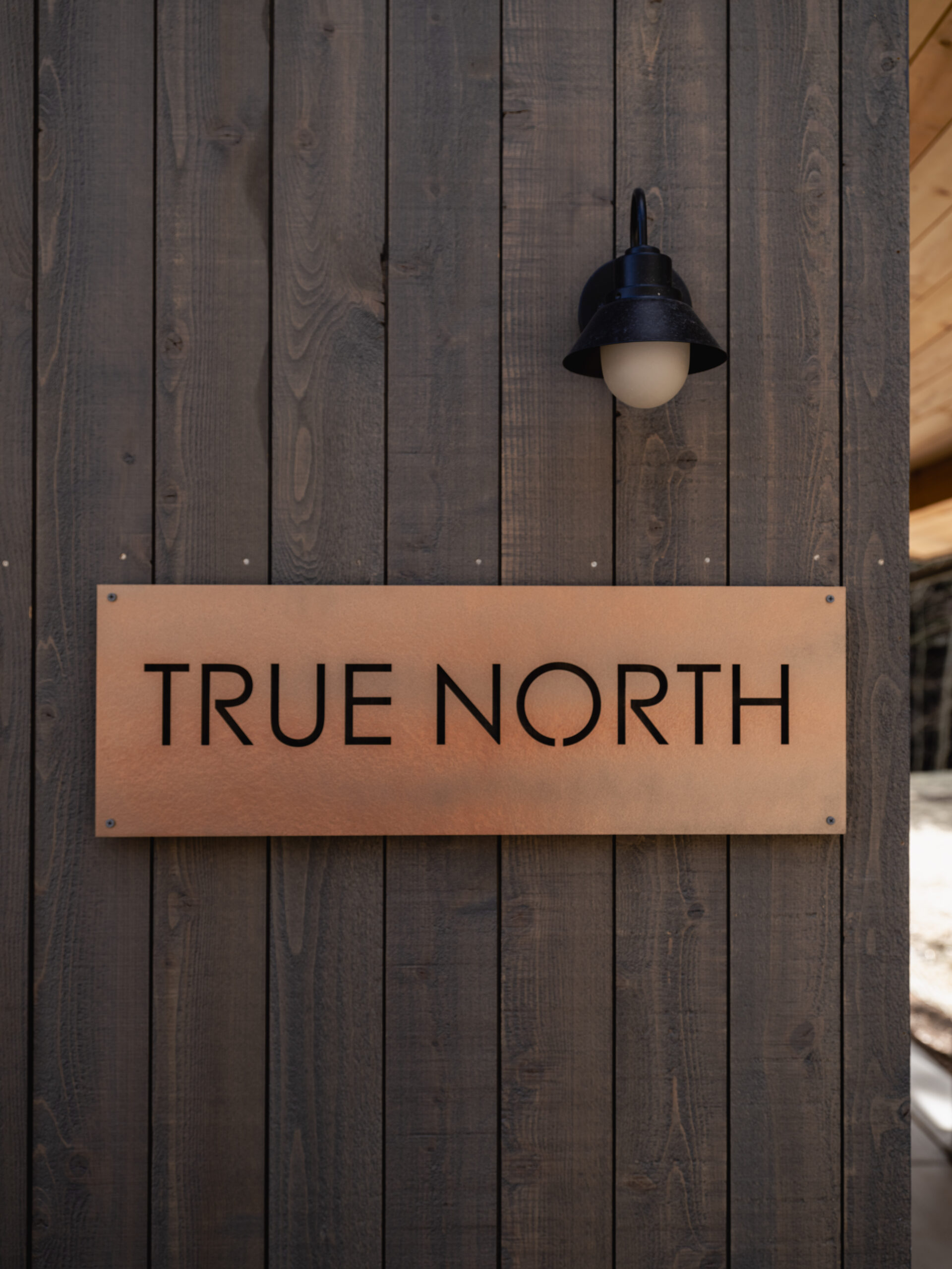 A copper sign reading "TRUE NORTH" is mounted on a dark wooden wall beneath a black outdoor wall lamp with a rounded white bulb.