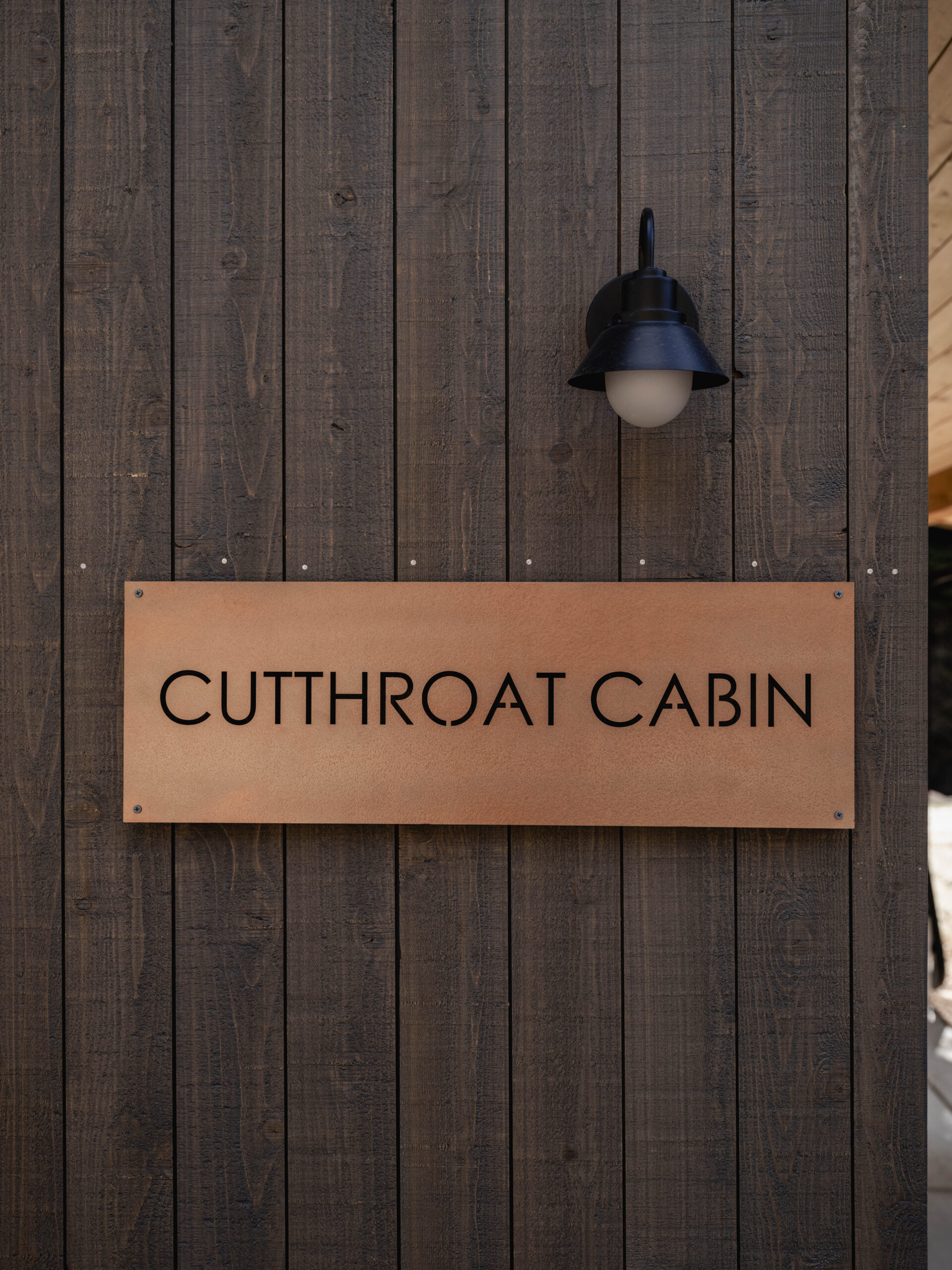 A wooden wall with a metal sign reading "CUTTHROAT CABIN" in all caps beneath a black outdoor light fixture.