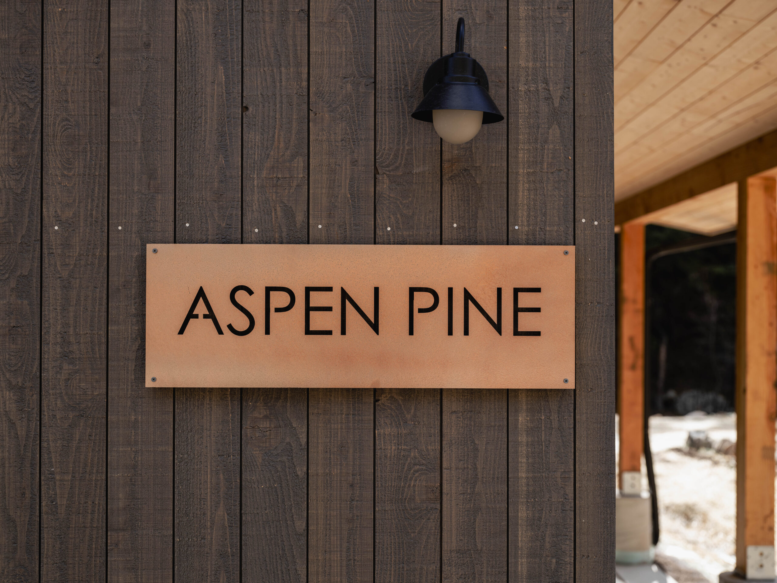 A wooden wall with a rectangular metal sign that reads "ASPEN PINE" in bold black letters, below an outdoor light fixture. Part of a wooden structure is visible to the right.
