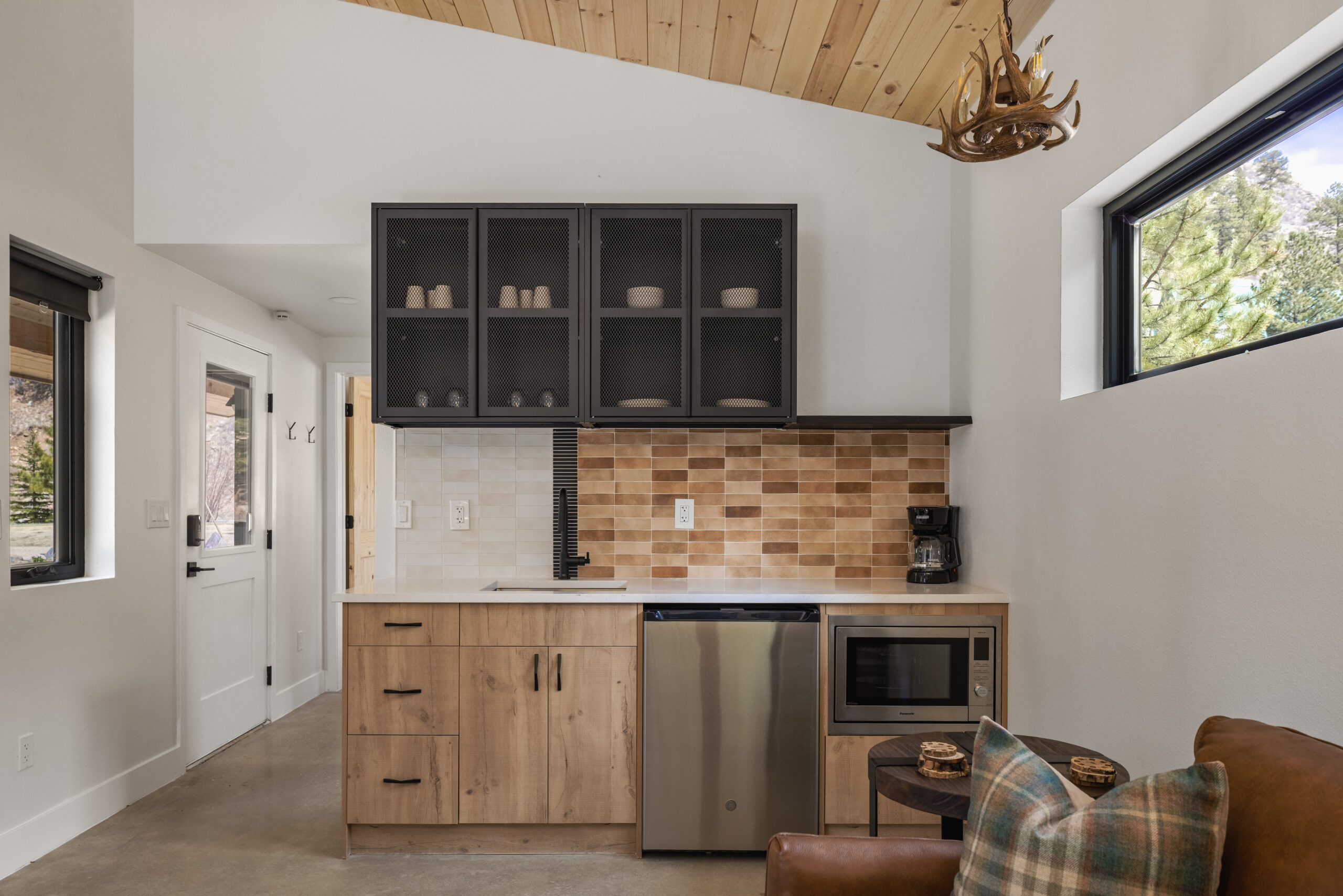 A small modern kitchenette with wooden cabinets, a mini fridge, a coffee maker, a tiled backsplash, glass-front upper cabinets, and a deer antler light fixture. A window and part of a brown leather sofa are visible.