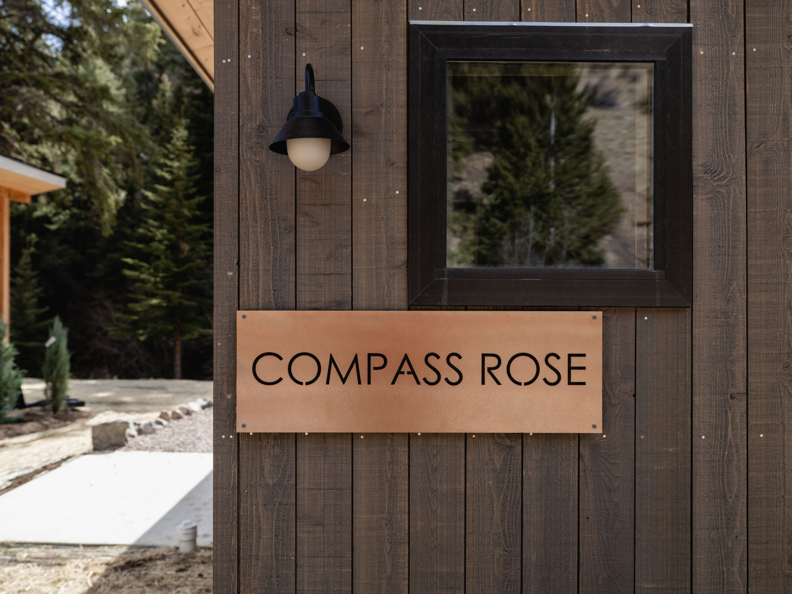A wooden building exterior with a black-framed window, a wall-mounted black lantern, and a rectangular sign reading "COMPASS ROSE" in large black letters. Trees and another building are visible in the background.