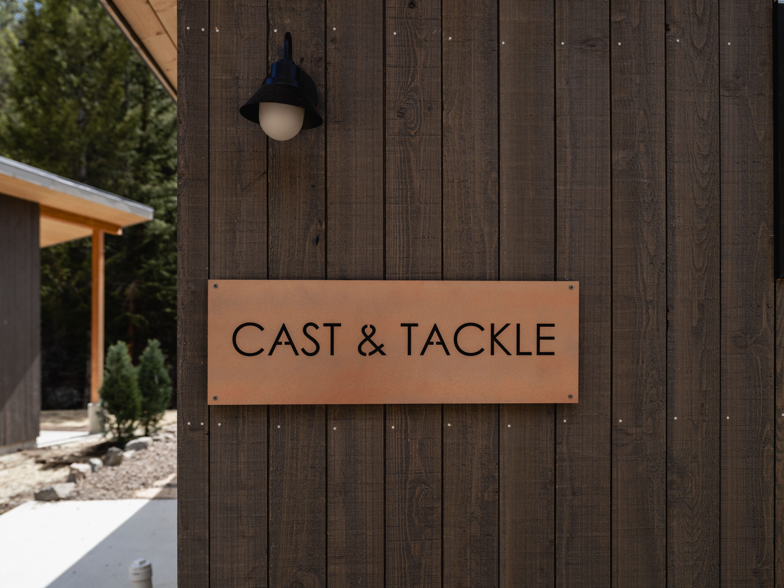 A wooden building exterior with a brown metal sign that reads "CAST & TACKLE" in black letters, mounted beneath a black outdoor light fixture. Trees and another building are in the background.