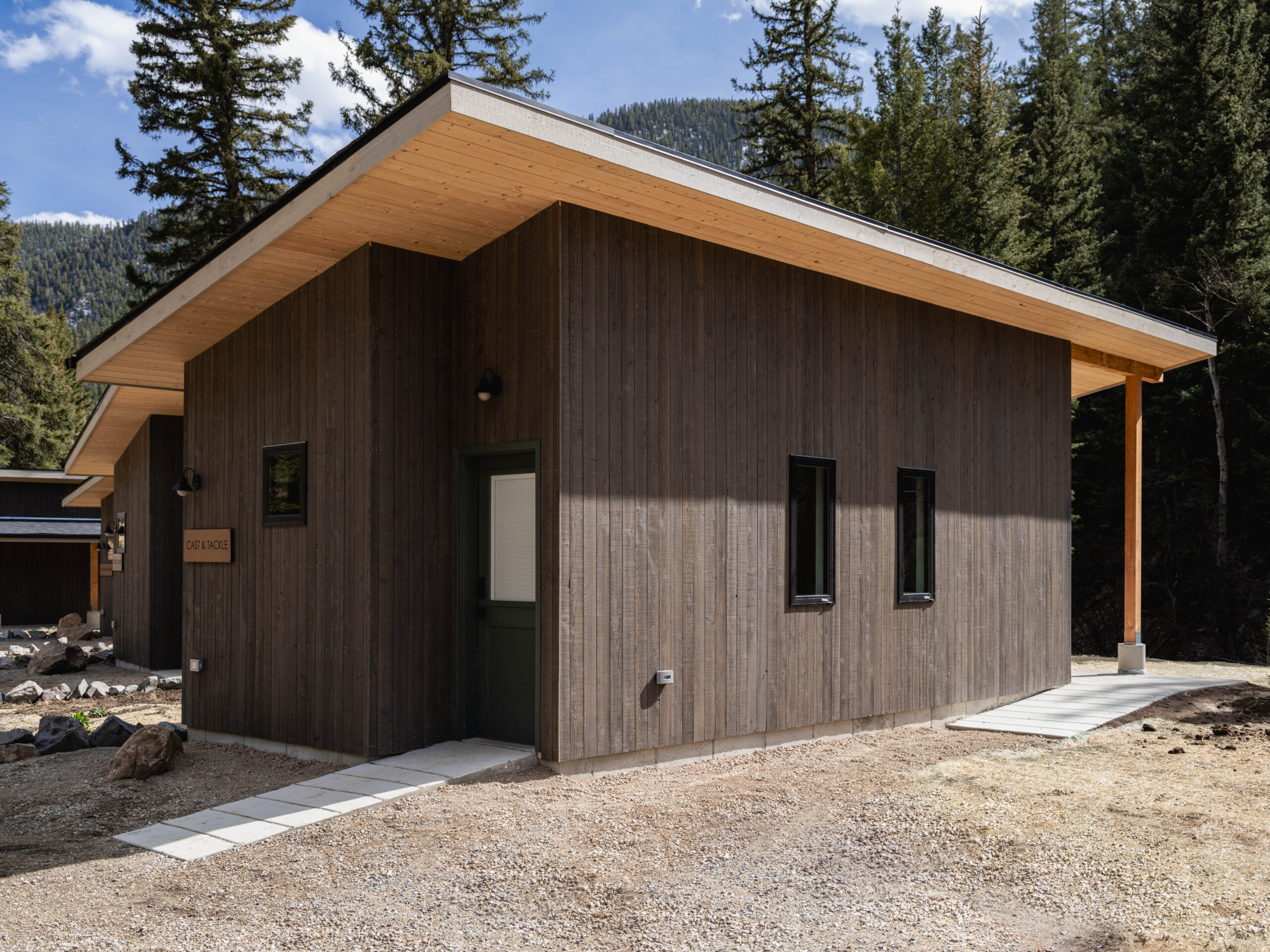 A modern cabin with dark wood siding, a sloped roof, and small rectangular windows, set among trees with mountains visible in the background. A pathway leads to the door.