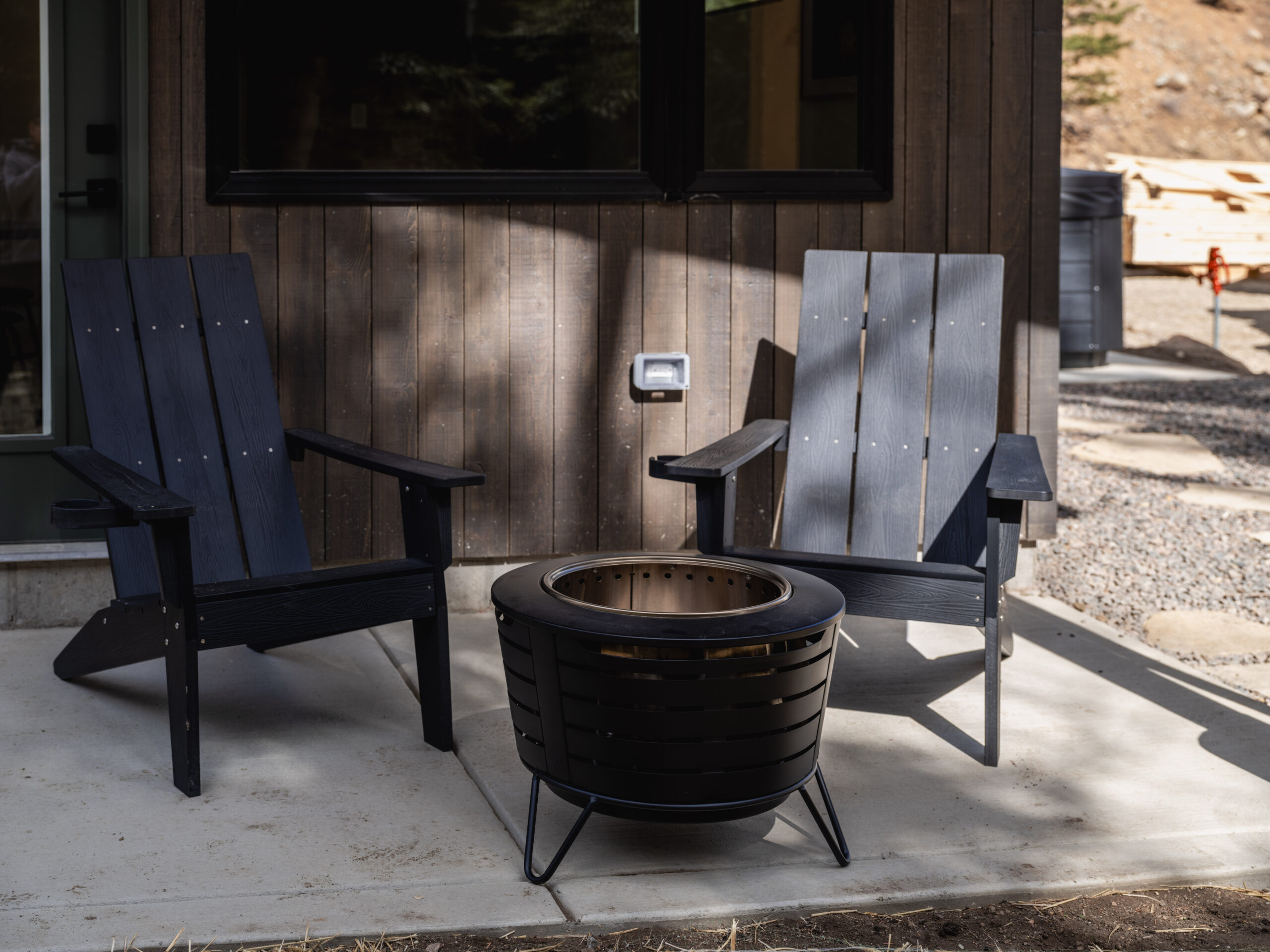 Two dark wooden Adirondack chairs sit on a concrete patio facing a modern black metal fire pit, outside a building with dark wood siding and a window. Gravel and trees are visible in the background.