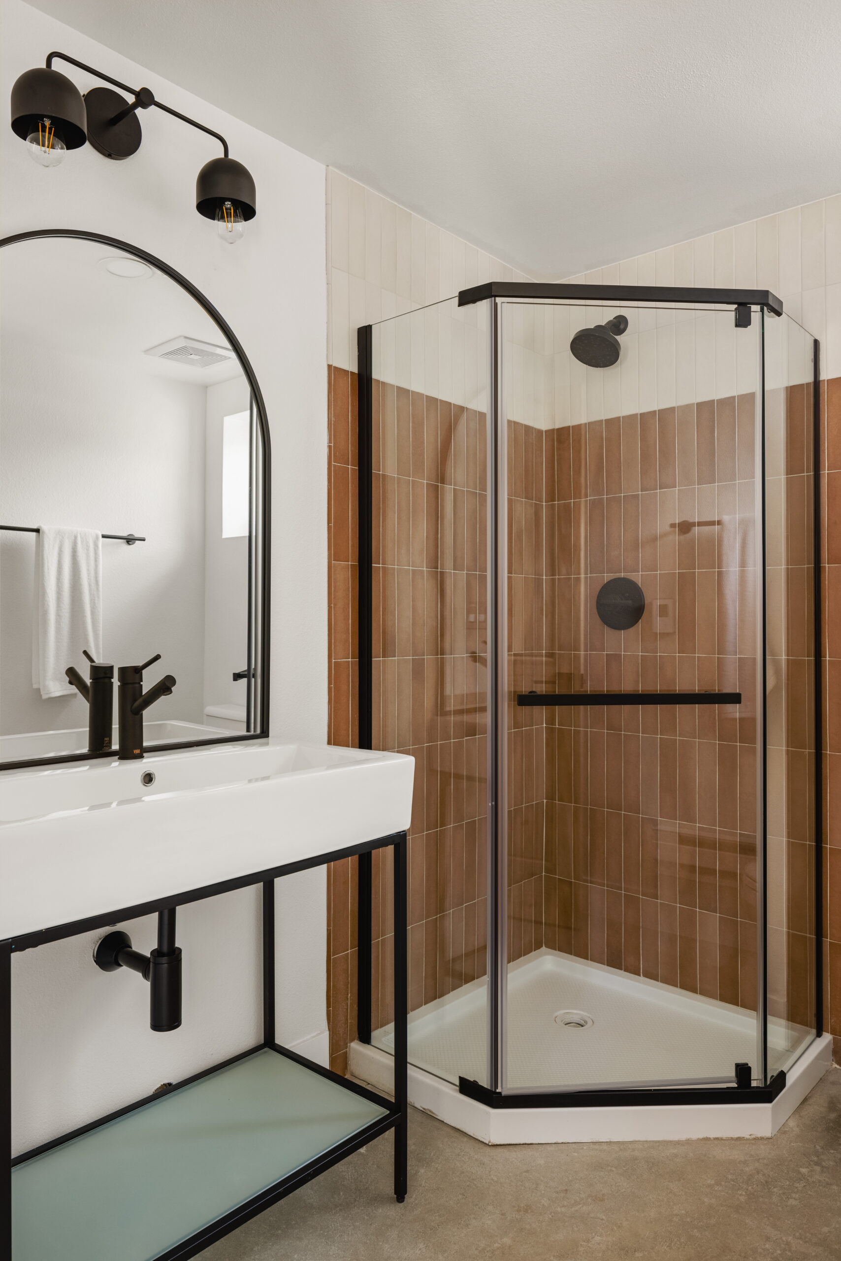 Modern bathroom with a glass-enclosed corner shower featuring beige tiles, a white rectangular sink with black fixtures, an arched mirror, wall-mounted lights, and a white towel hanging on the wall in the background.