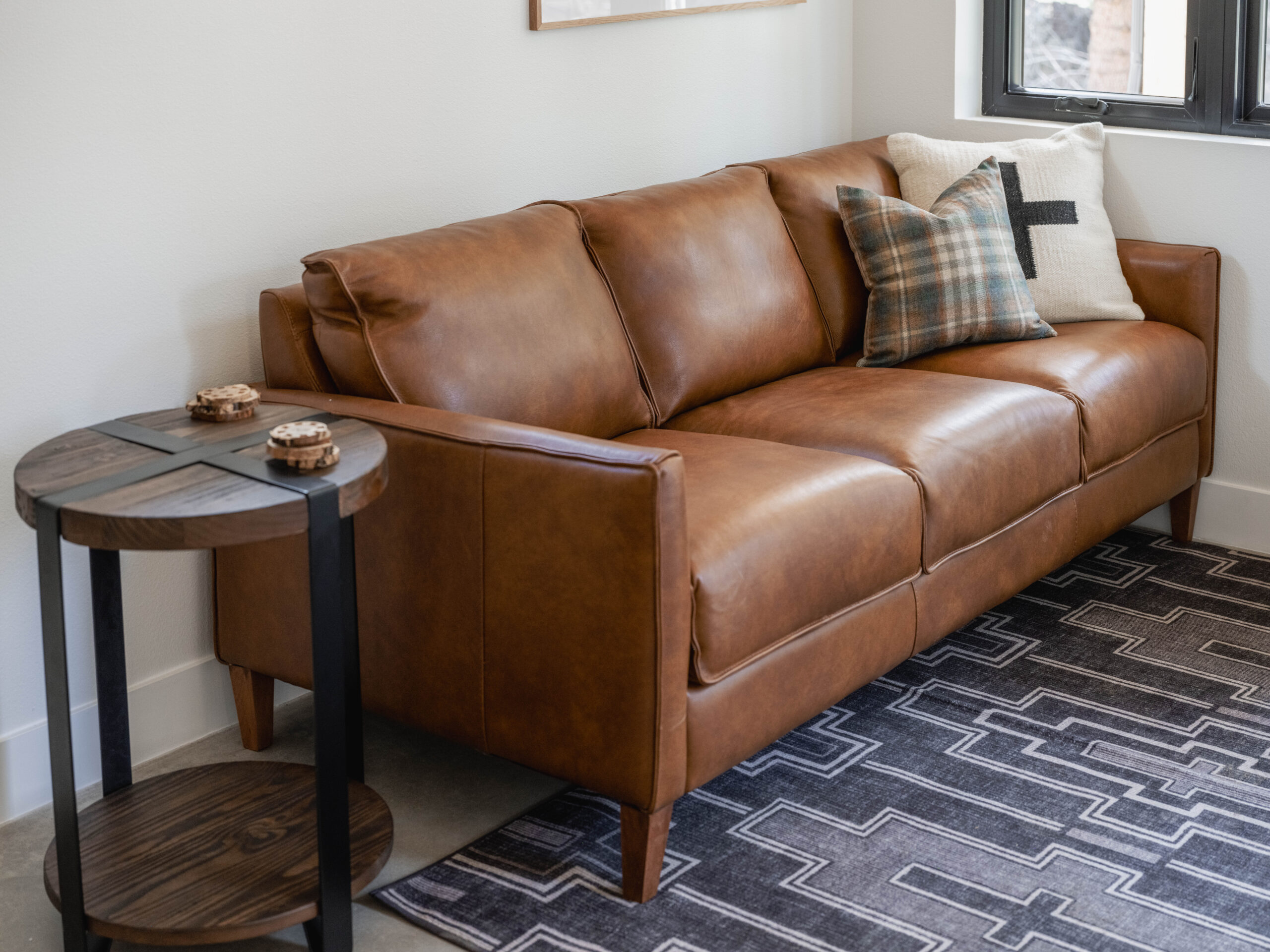 A brown leather sofa with three cushions sits on a patterned rug next to a round wooden side table holding two small objects, in a modern living room with light walls and a window.