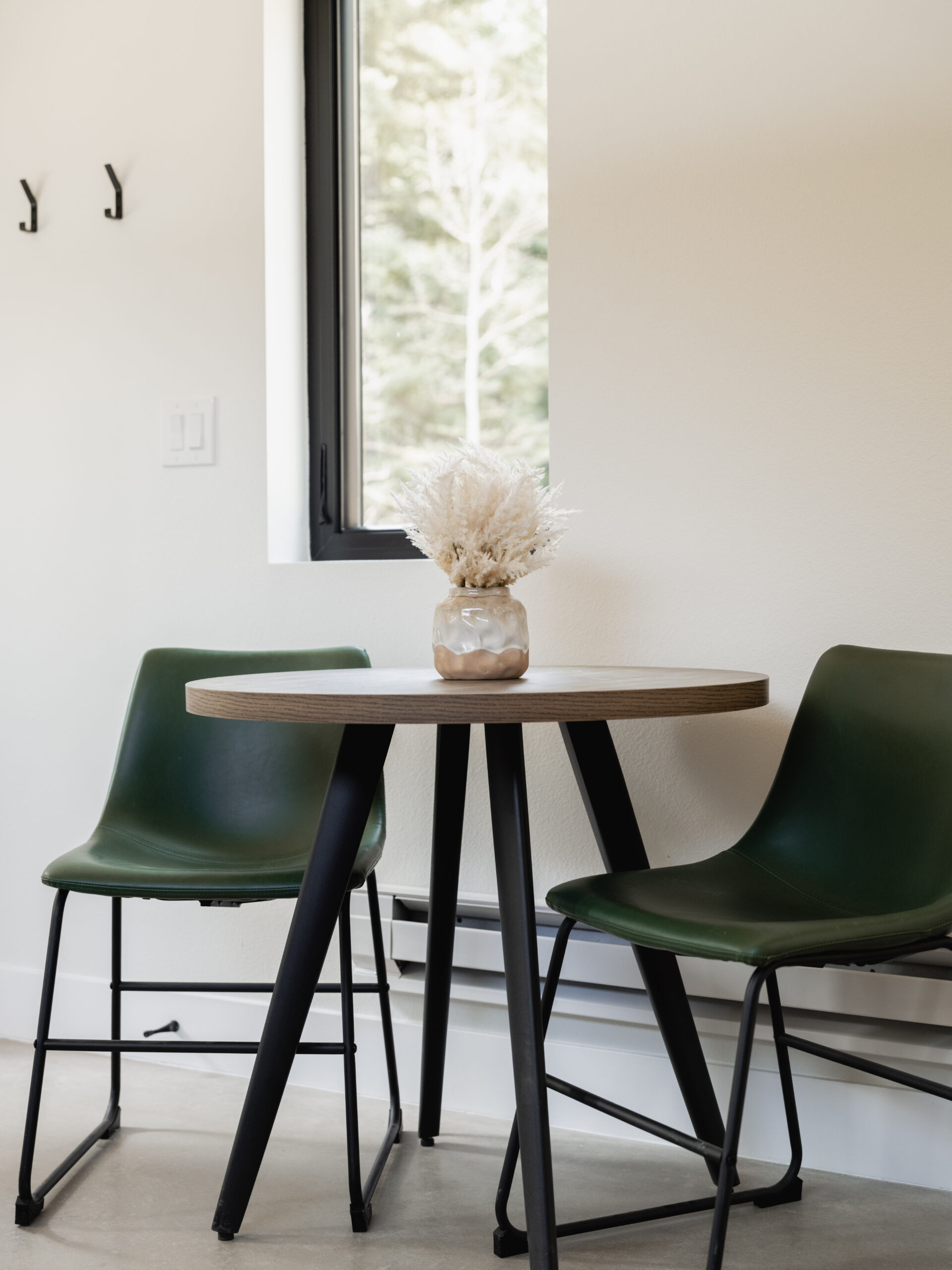 A small round table with a light wood top and black legs sits between two dark green chairs. A glass jar with dried white flowers is centered on the table. A window and white wall are in the background.