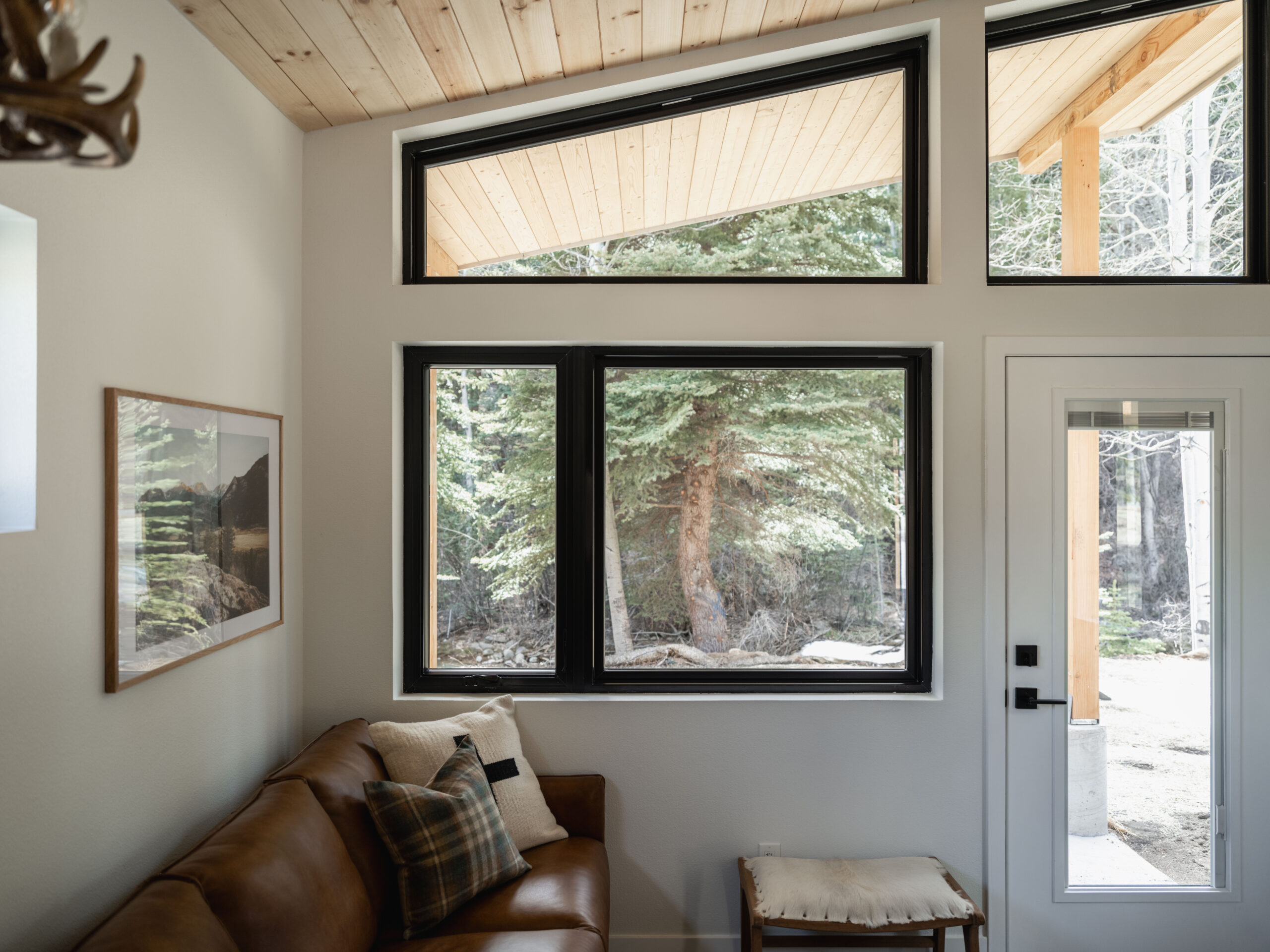 A cozy room with a brown leather sofa, plaid and beige pillows, a small cushioned stool, and large windows framing a view of pine trees and a wooden patio outside. Light wood ceiling and walls create a warm atmosphere.