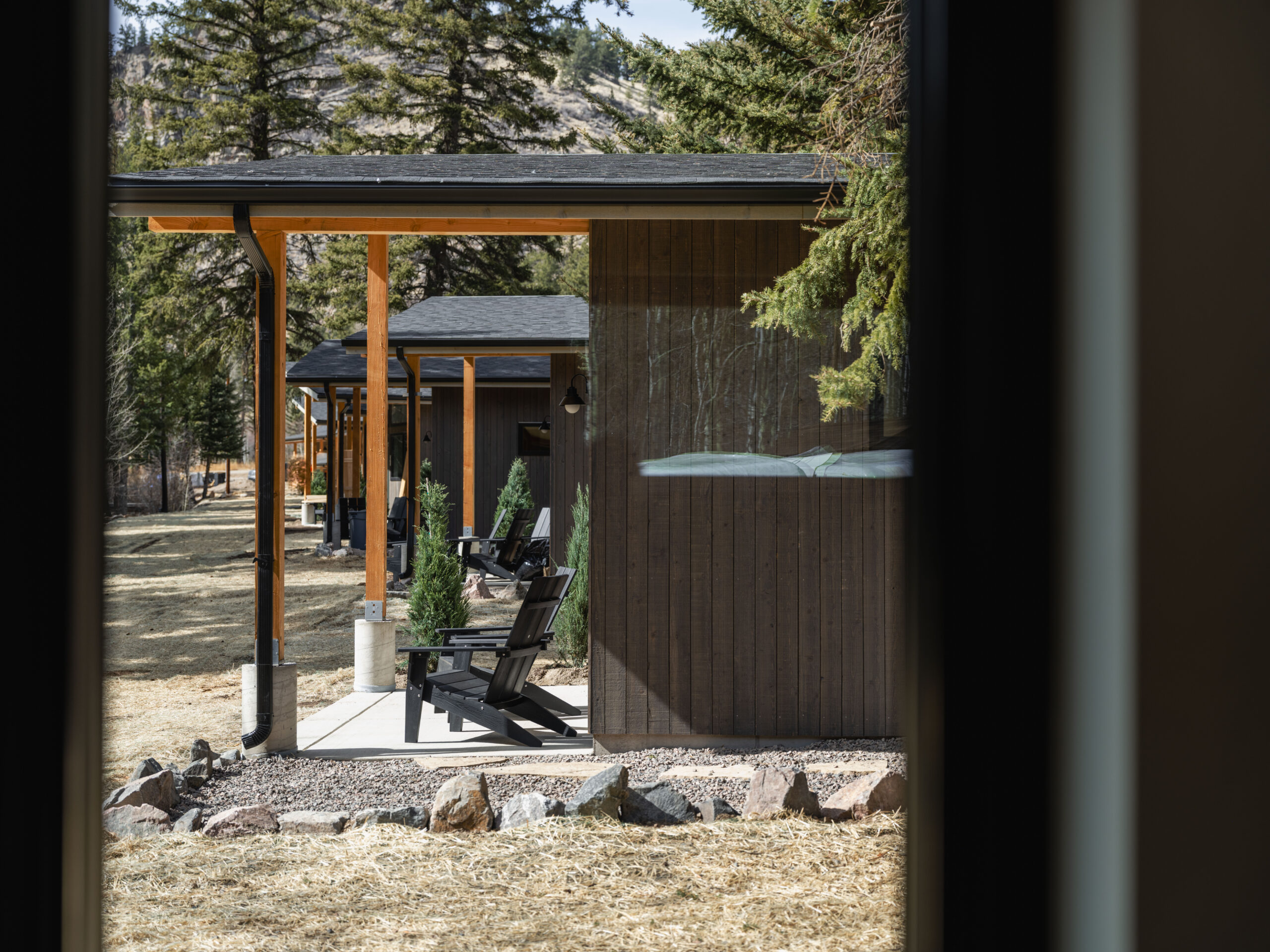 View through a window of a modern wooden cabin with a small patio, black chairs, and trees in the background, surrounded by nature and natural landscaping.