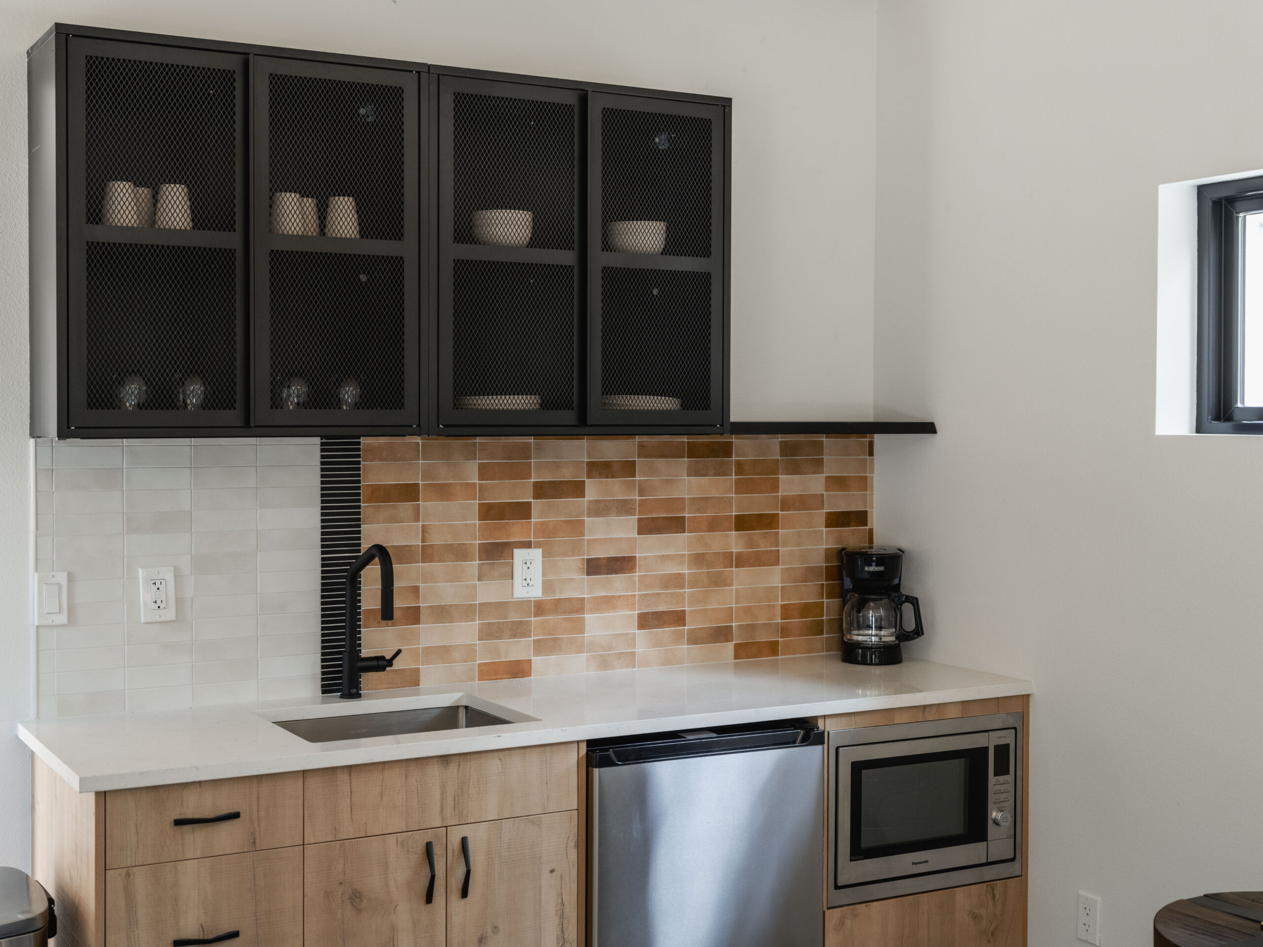 A modern kitchen with light wood cabinets, a small sink, black faucet, dishwasher, microwave, coffee maker, and upper cabinets with mesh doors. The backsplash is tiled in white and brown tones.