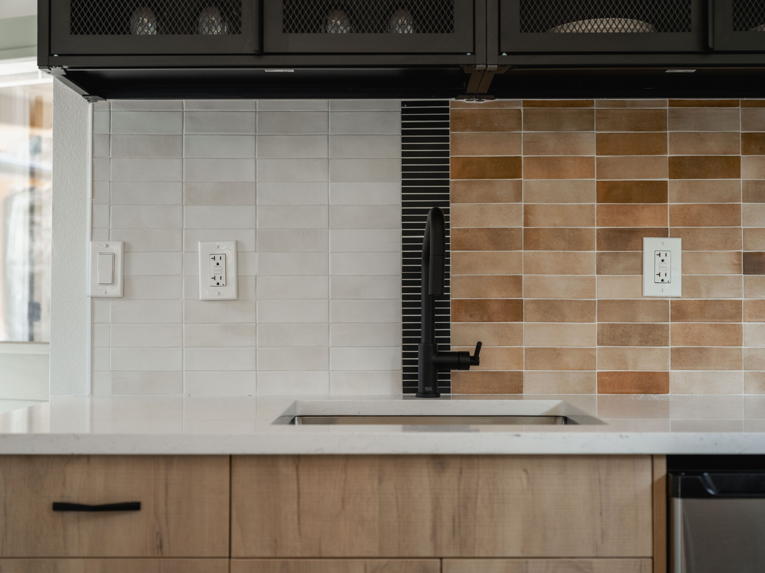 A modern kitchen backsplash divided in two styles: white subway tiles on the left and brown tiles on the right. A black faucet and sink are centered, with light wood cabinets below and dark cabinets above.