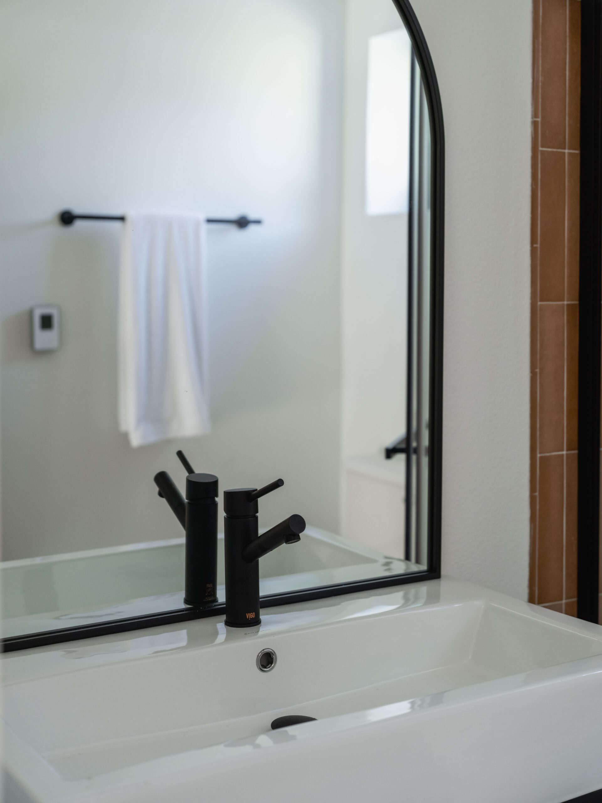 A modern bathroom sink with a black faucet is reflected in a large mirror. A white towel hangs on a black towel bar on the wall, and natural light brightens the minimalist space.