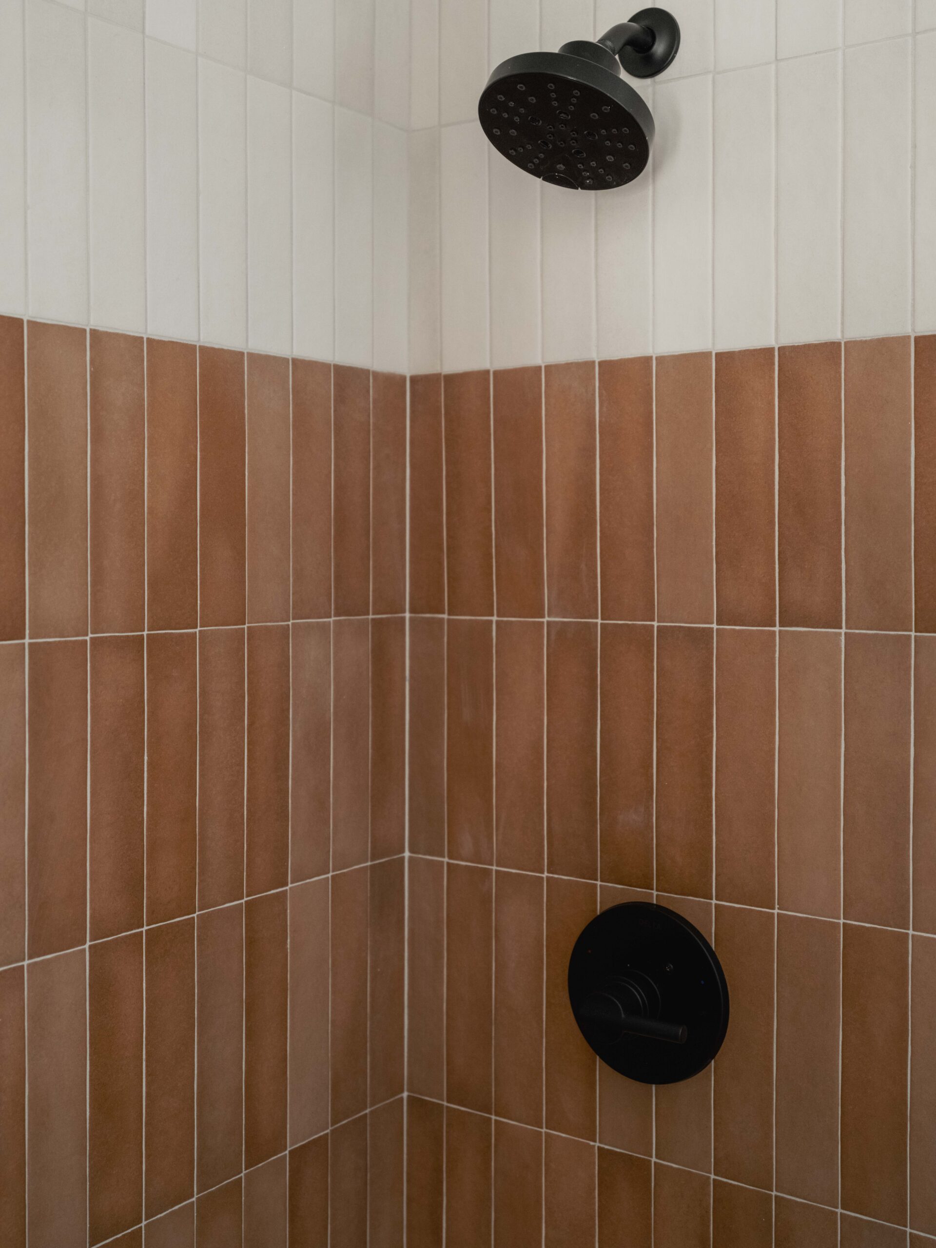 A shower with vertical rectangular tiles in two colors: white on the upper half and brown on the lower half. It has a black showerhead and a matching black control knob.