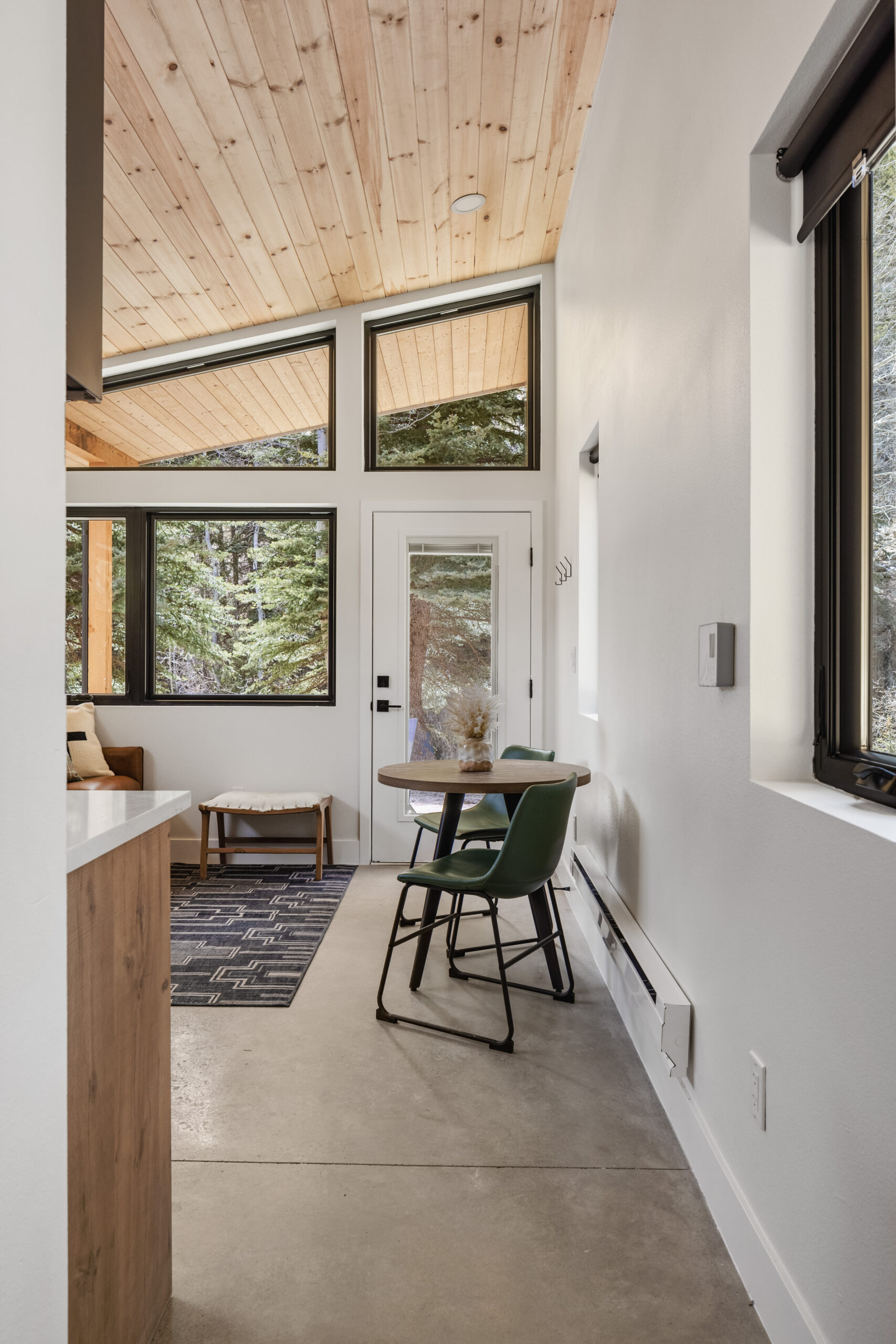 Small modern dining area with two green chairs at a round table, large windows, a wooden ceiling, and a door leading outside to a forested area. Natural light fills the room.