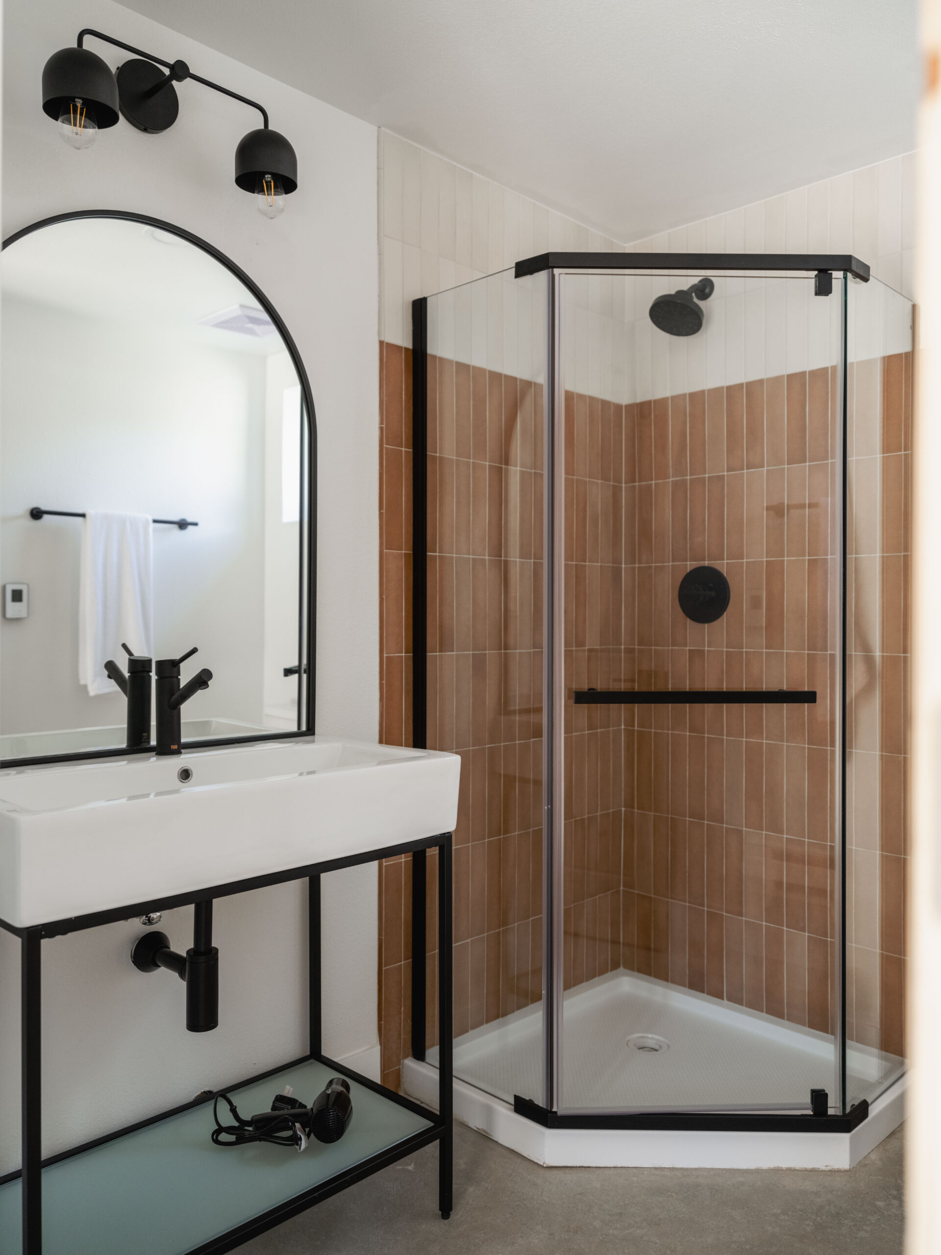 Modern bathroom with a corner glass shower featuring tan tiles, a white sink with a black faucet and frame, an arched mirror, black wall lights, and a towel hanging in the background.