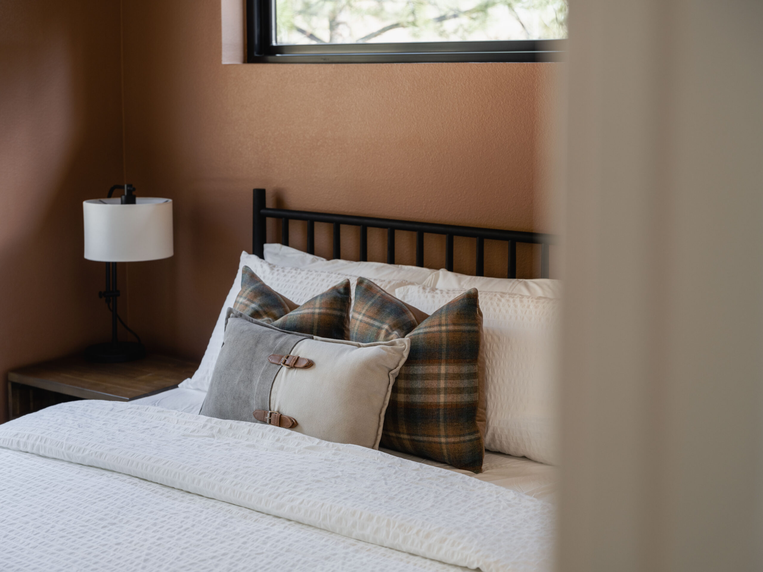 A neatly made bed with white bedding, plaid and solid throw pillows, a black metal headboard, and a bedside table with a lamp, set against a brown wall with a window above.
