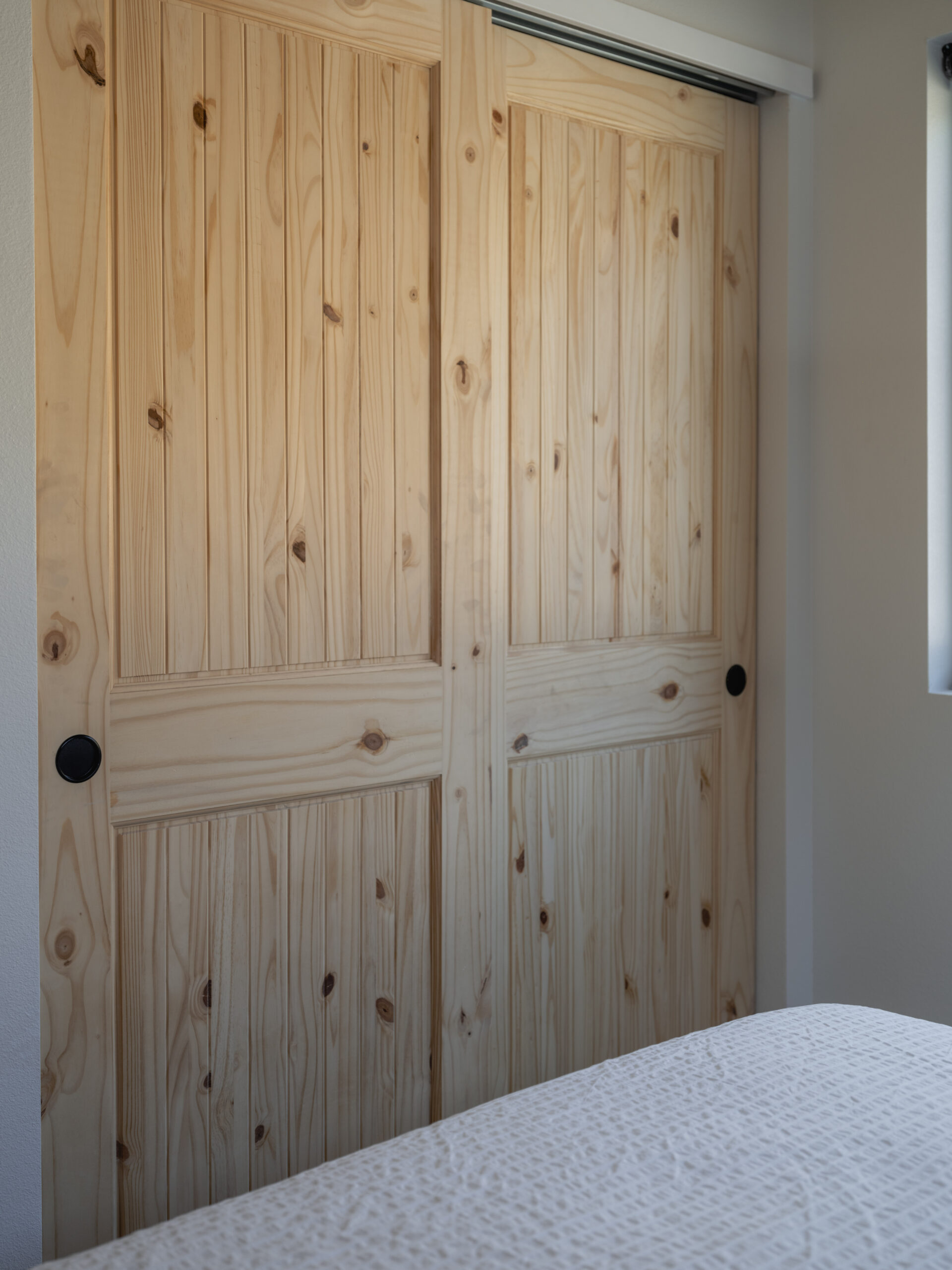 Two light wood sliding closet doors with visible knots and grain, closed, with round black handles. Part of a bed with a white textured bedspread is visible in the foreground. Light enters from a window on the right.