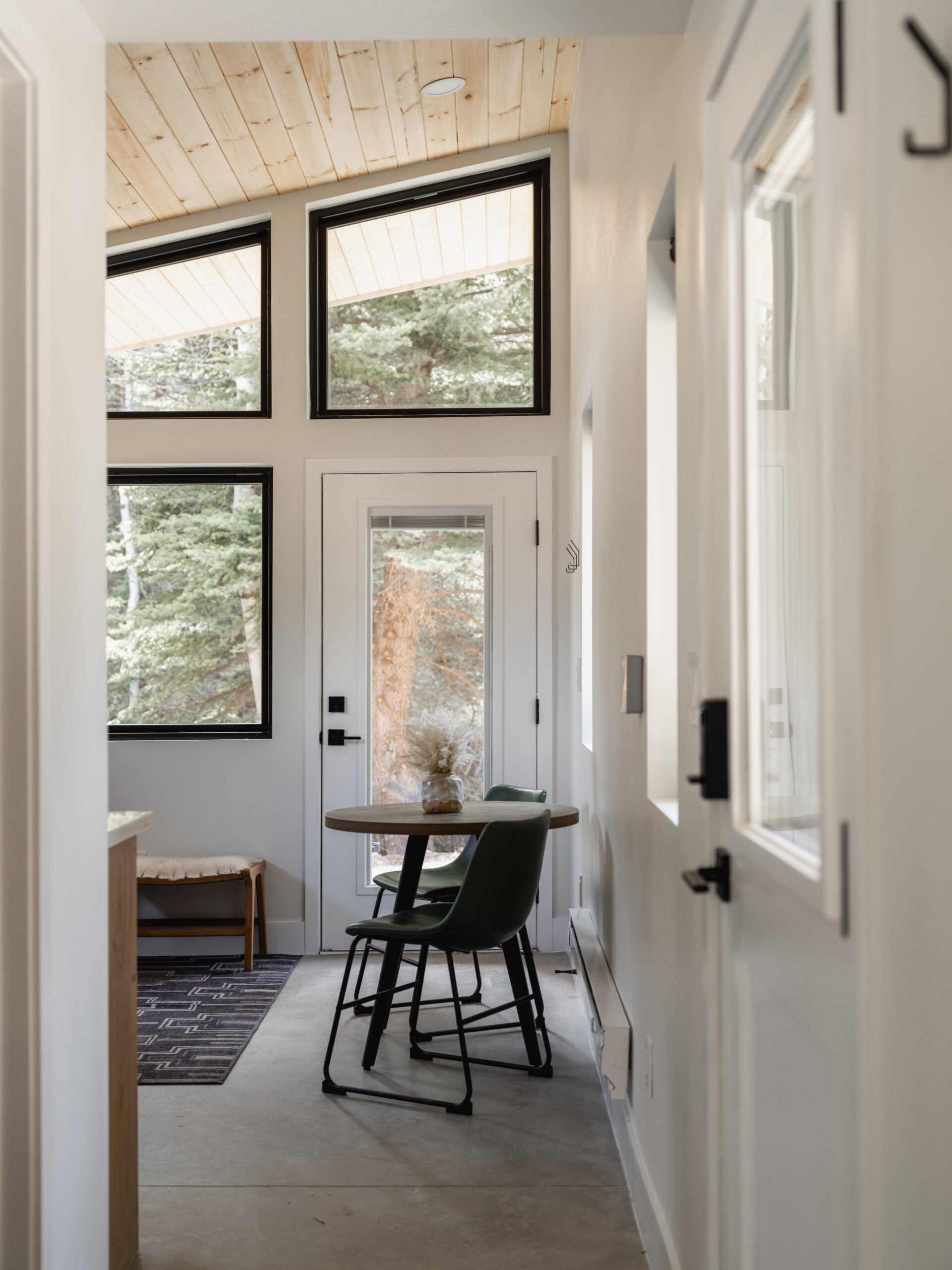 Small, modern entryway with large windows, a round table with two green chairs, a bench, and views of trees outside. Light wood ceiling and white walls create a bright, airy atmosphere.