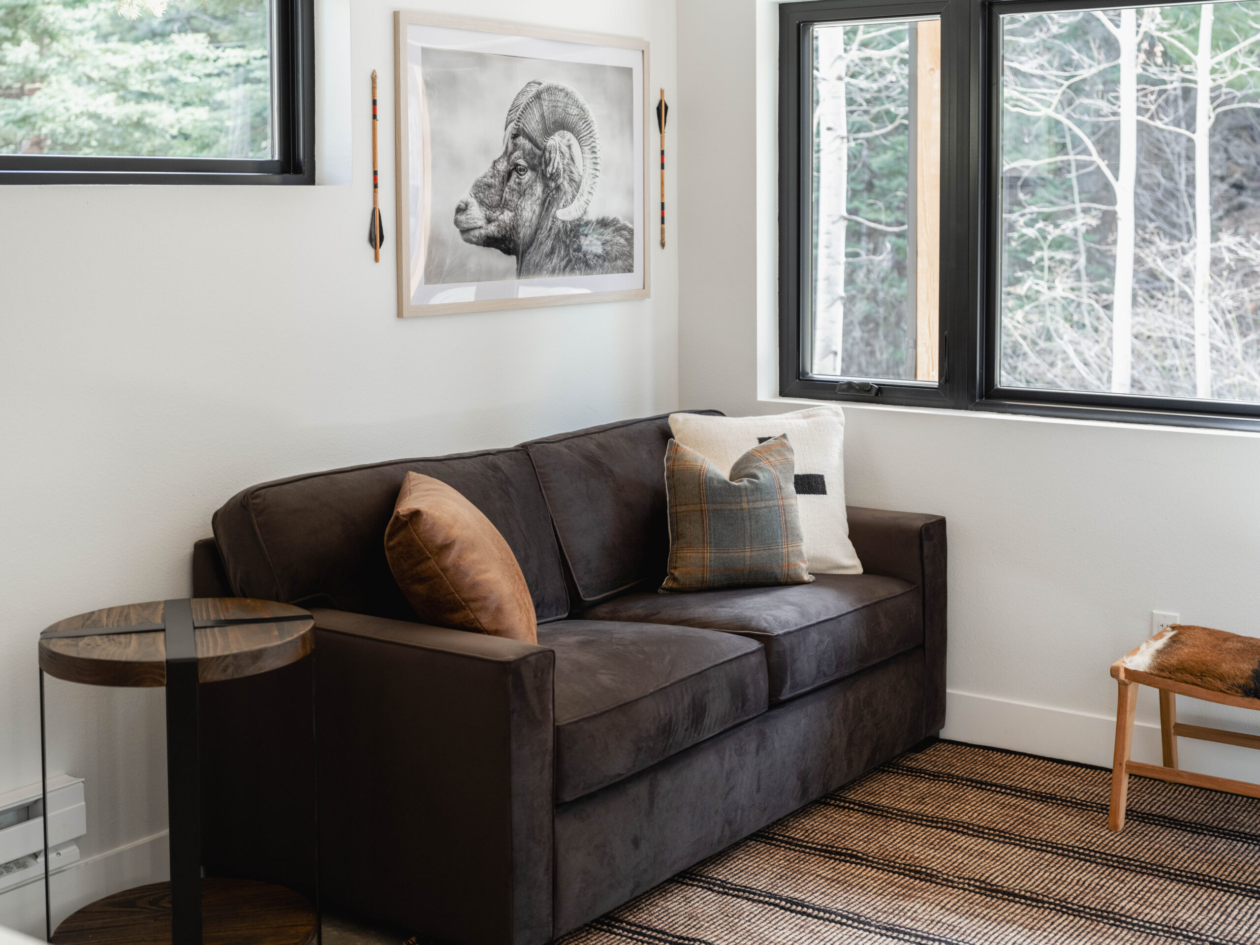 A dark brown sofa with two decorative pillows sits against a white wall, topped by a framed ram illustration. A round glass-topped side table and a window with a view of trees complete the cozy, modern room.