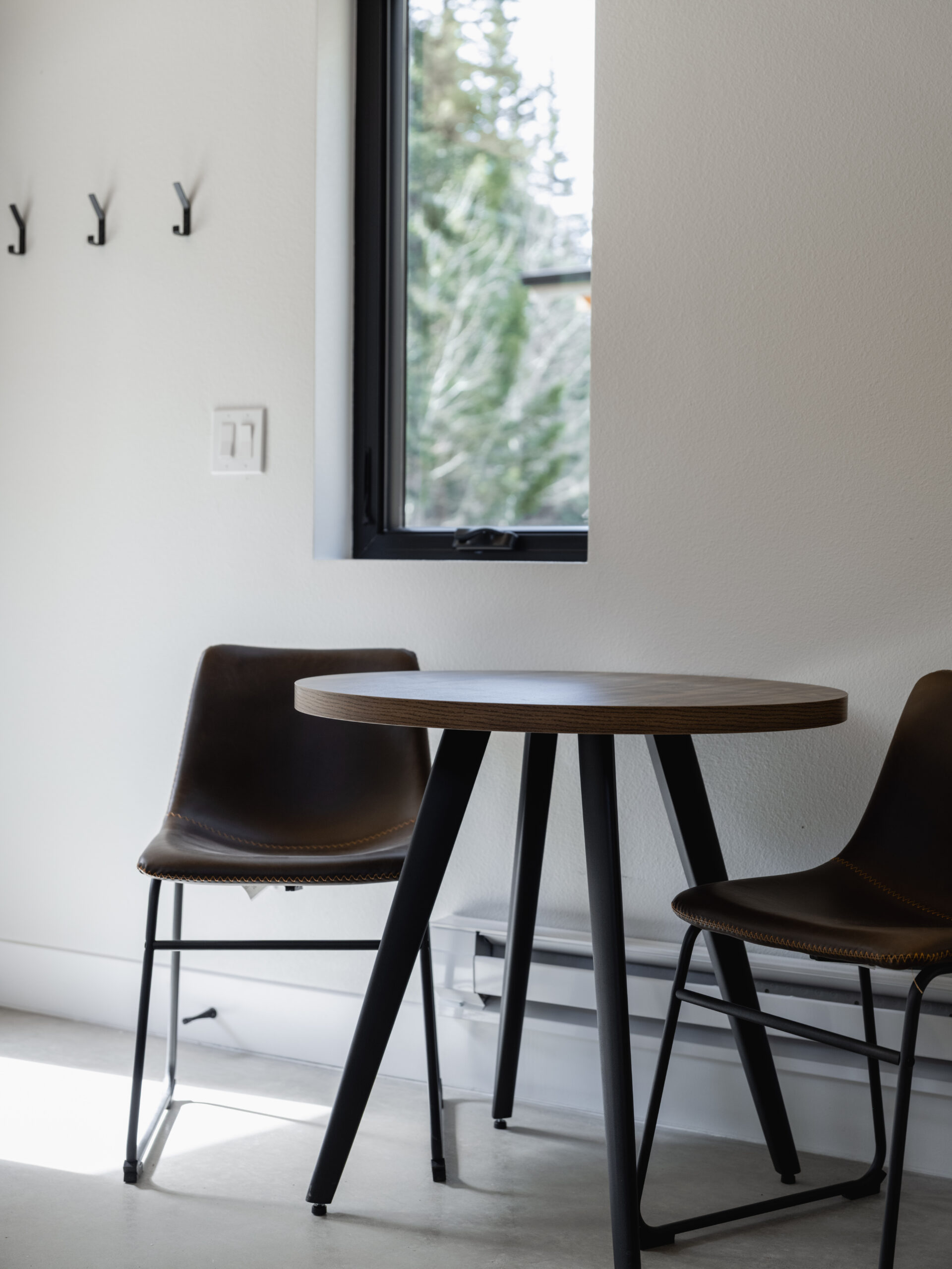 A small round table with two dark chairs is placed near a window in a bright, minimalist room. Wall hooks are visible to the left, and trees can be seen through the window.