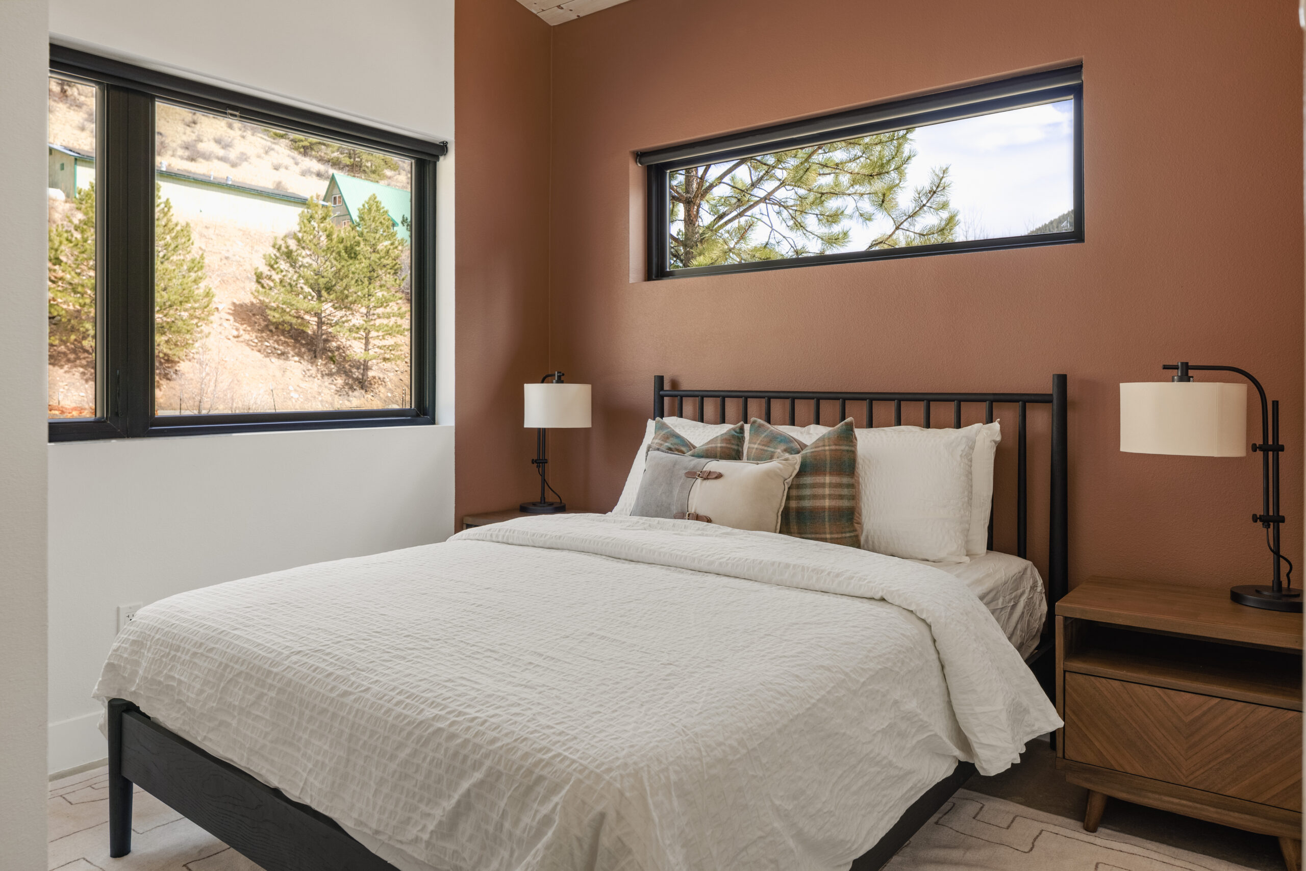 A cozy bedroom with a black metal bed frame, white bedding, plaid and neutral pillows, two wooden nightstands with lamps, and windows showing trees and hills outside. The walls are white and brown.