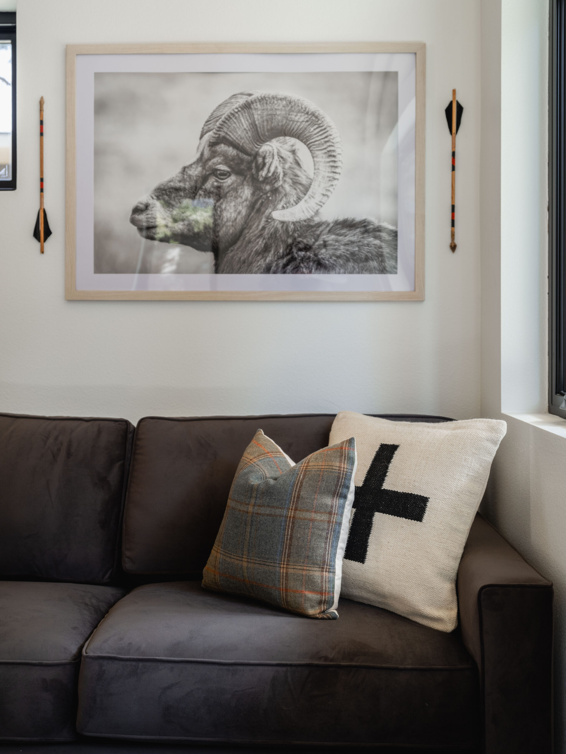 A black and white framed photo of a ram hangs above a dark gray sofa with two pillows, one plaid and one with a black cross. Two decorative arrows are mounted on the wall beside the artwork.