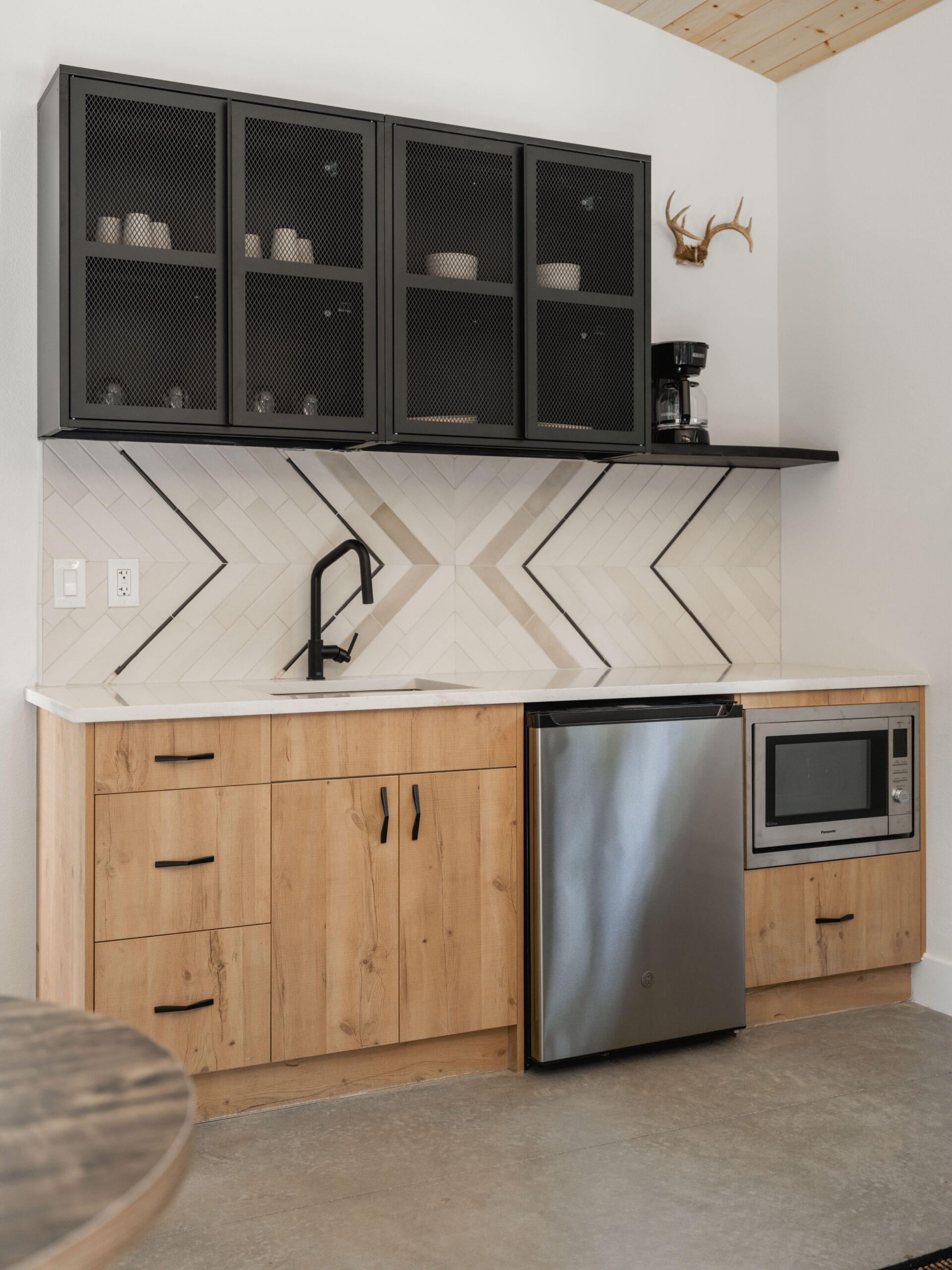 Modern kitchenette with light wood cabinets, a small stainless steel fridge, built-in microwave, black sink and faucet, black mesh upper cabinets, and chevron-patterned white tile backsplash. Antler decor on the wall.