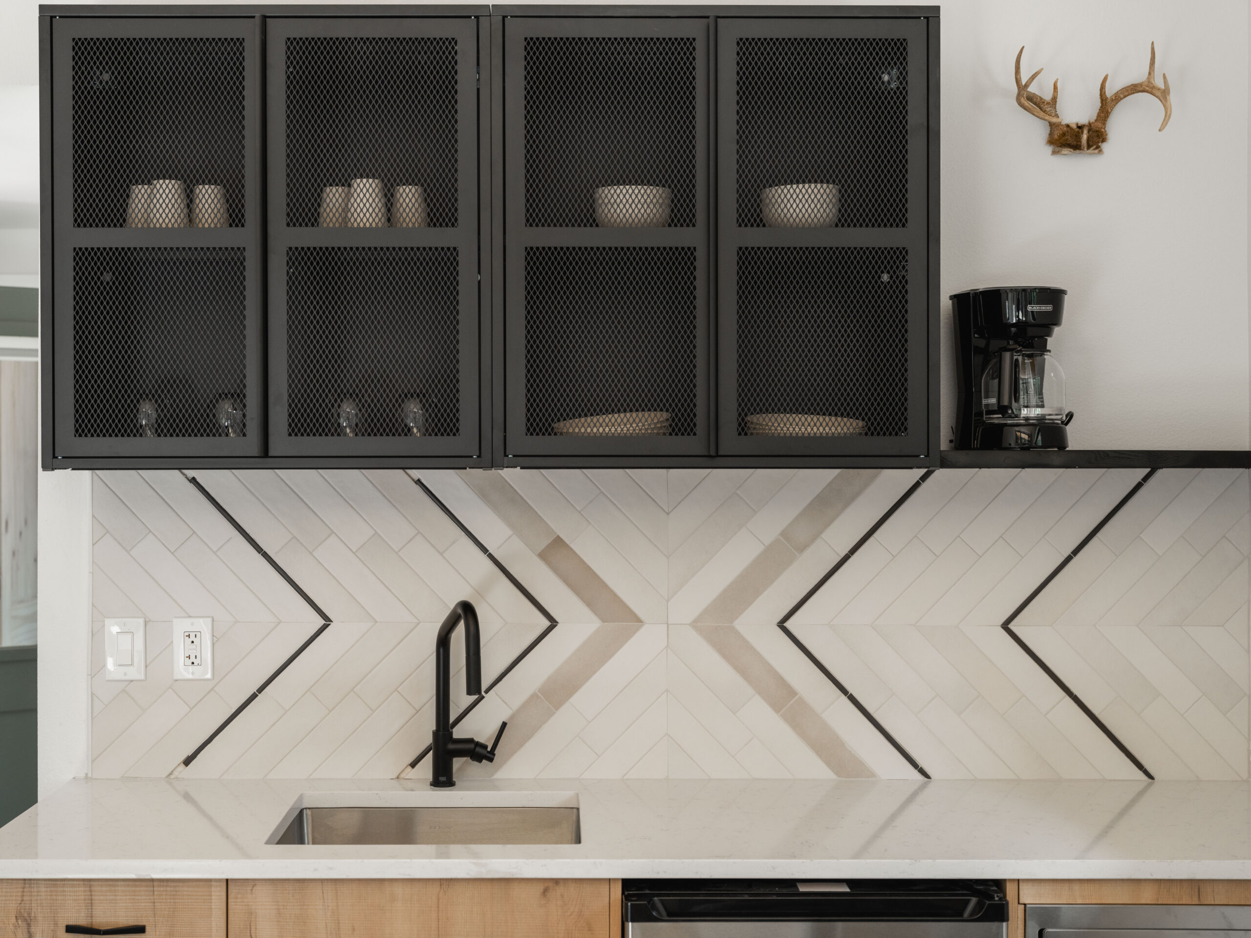 A modern kitchen features a black faucet, chevron-patterned white tile backsplash, black mesh upper cabinets with dishes, a coffee maker, and a decorative antler mount on the wall.