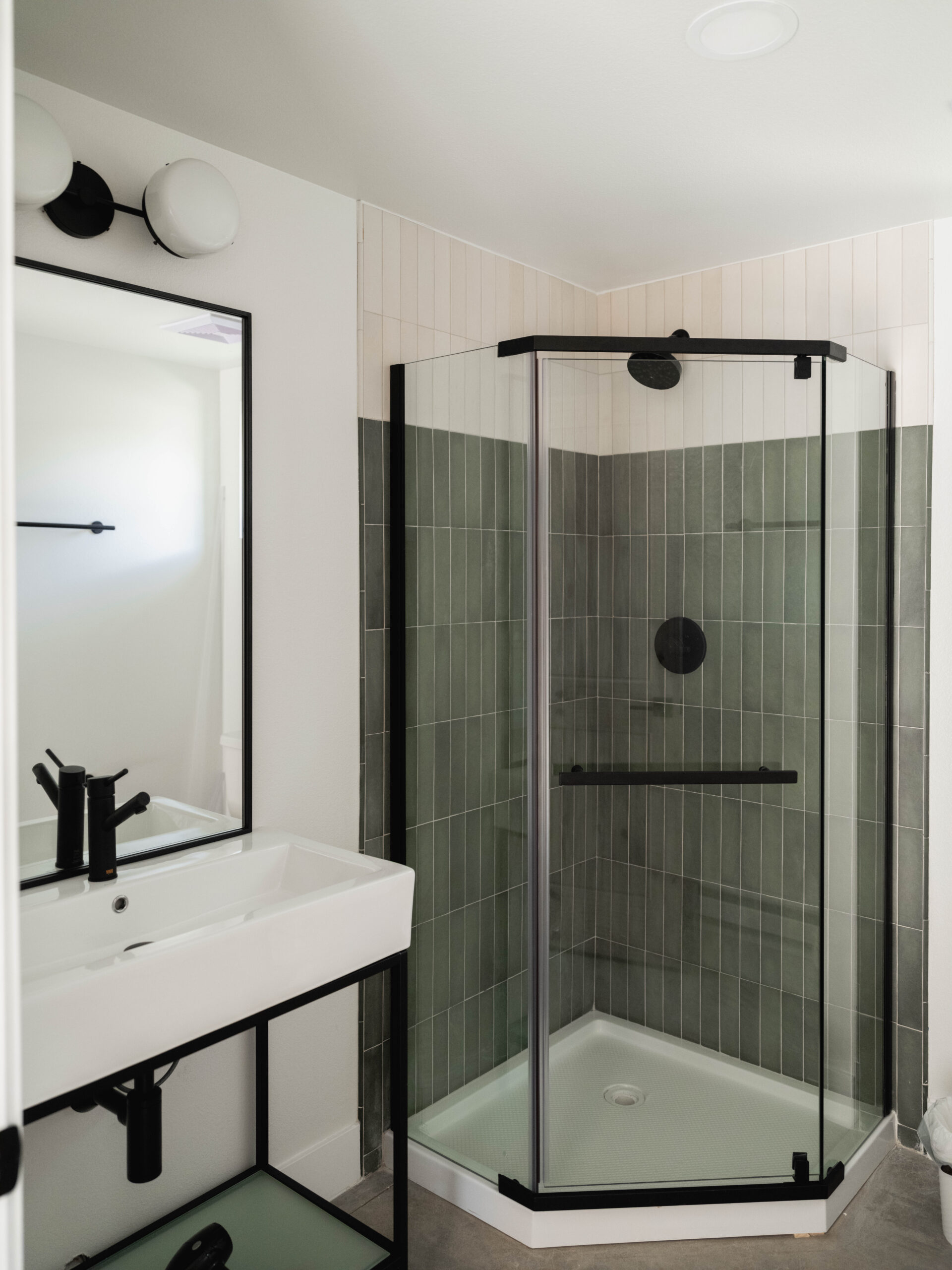 Modern bathroom with a corner glass shower enclosure, green vertical tiles, a rectangular white sink with black fixtures, a black-framed mirror, and a wall-mounted light above the mirror.