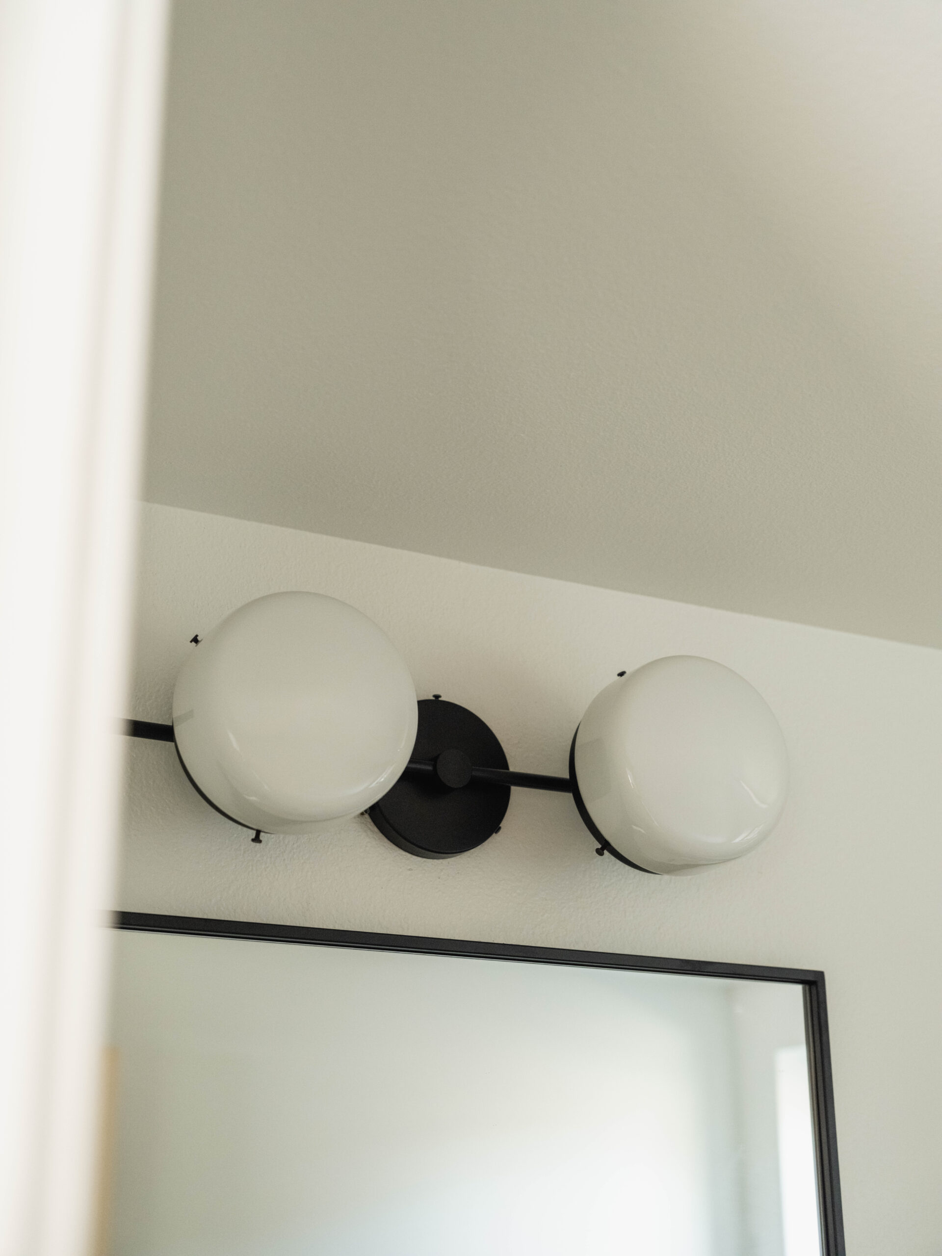 A close-up of a modern wall-mounted light fixture with two round, white glass globes and a black metal base, positioned above a rectangular mirror in a bathroom.