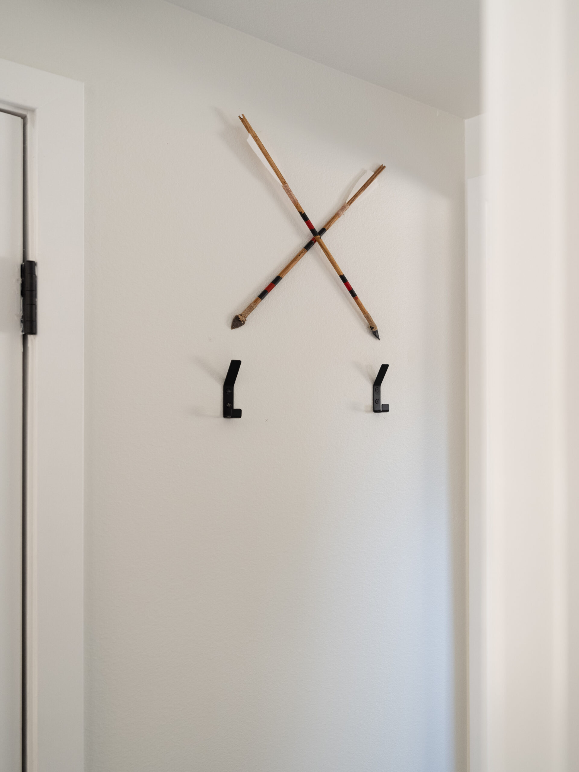 Two black wall hooks are mounted on a white wall beneath two crossed decorative wooden arrows. The space is minimal with a door frame visible on the left side.