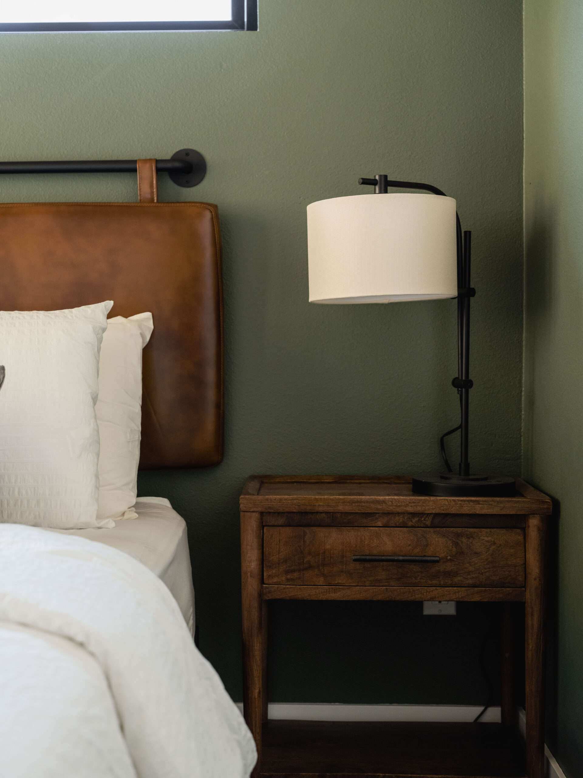 A wooden nightstand with a drawer and a modern black lamp sits next to a bed with white bedding and a brown leather headboard against a green wall.