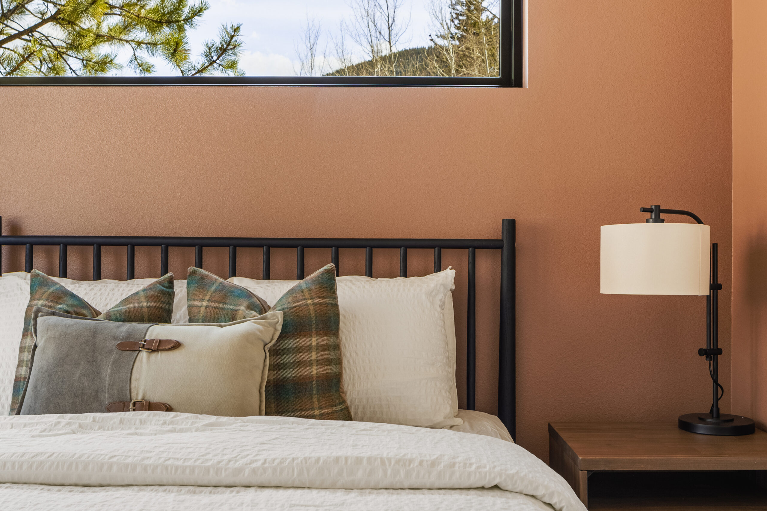 A cozy bed with plaid and solid pillows, a white comforter, and a black metal headboard sits beside a wooden nightstand with a modern lamp; a window above lets in natural light.