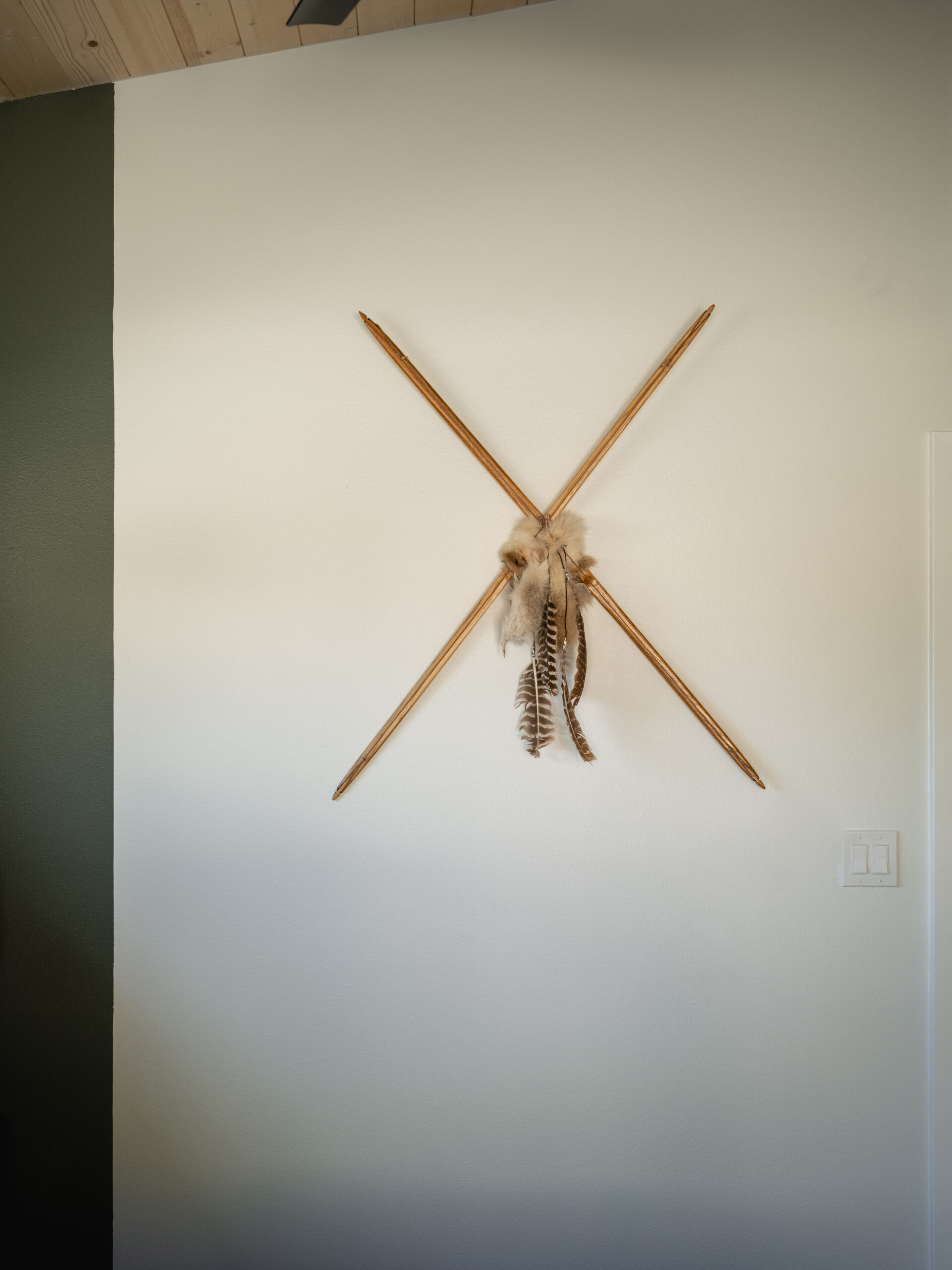 Two wooden spears arranged in an X shape hang on a white wall, decorated at the intersection with feathers and fur. The wall is partially green, and a light switch is visible to the right.