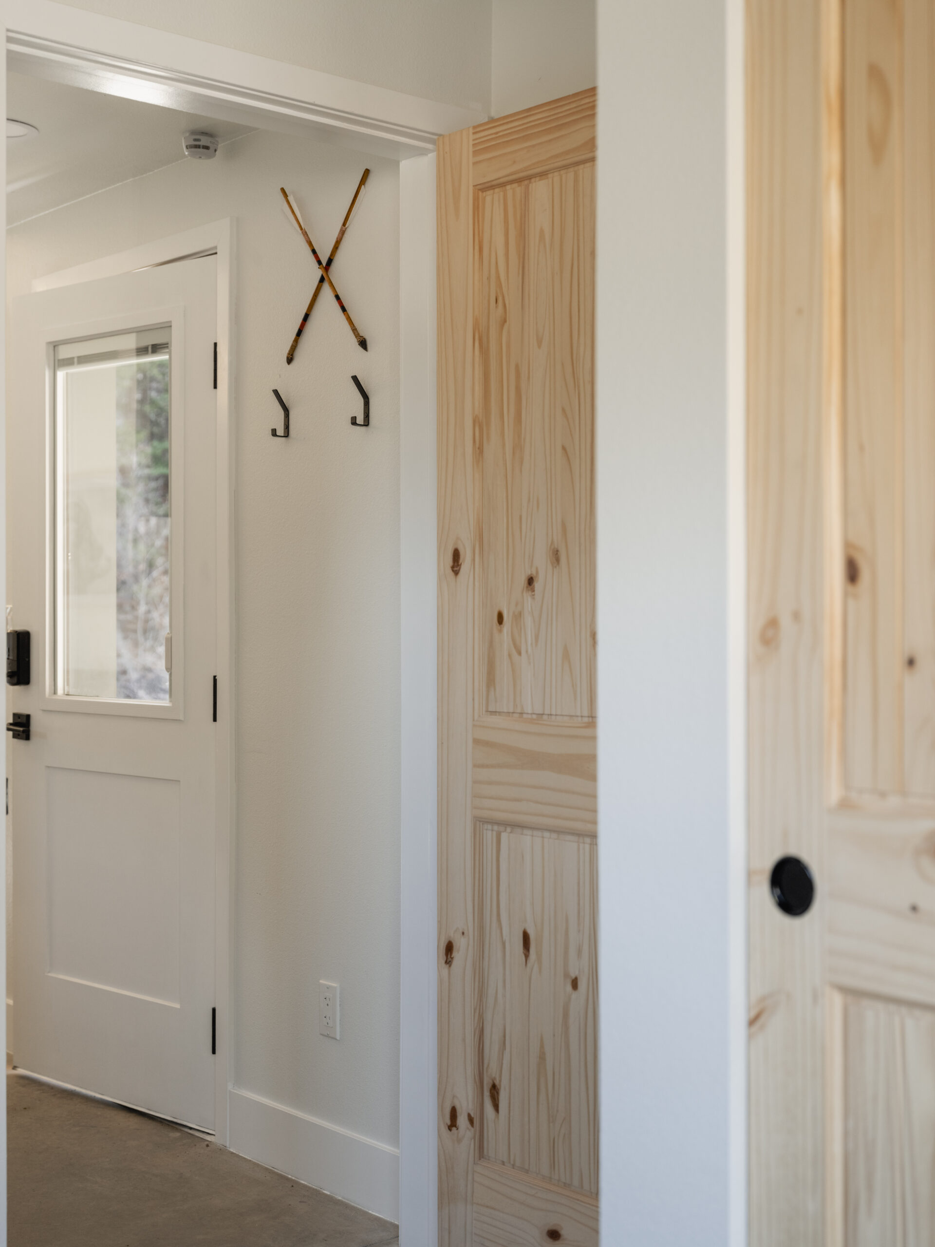 A minimalist entryway with a white door, a wooden closet door, and two wall hooks shaped like an X, set against light-colored walls. Natural light brightens the space.