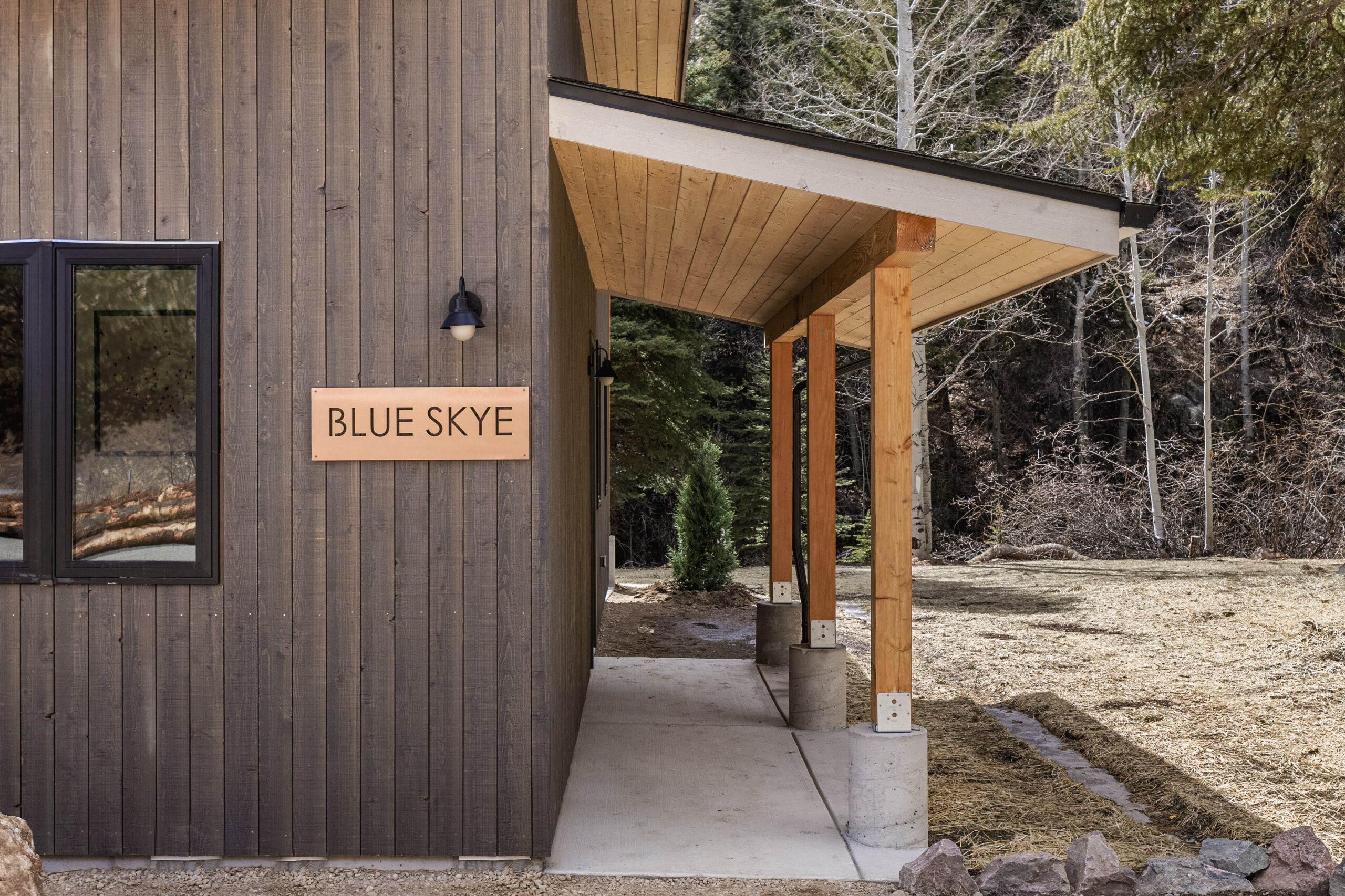 A wooden building with a sign reading "BLUE SKYE" mounted on the dark vertical siding. The building has a slanted roof, a window, and a covered walkway with wooden posts. Trees and a grassy area are visible in the background.