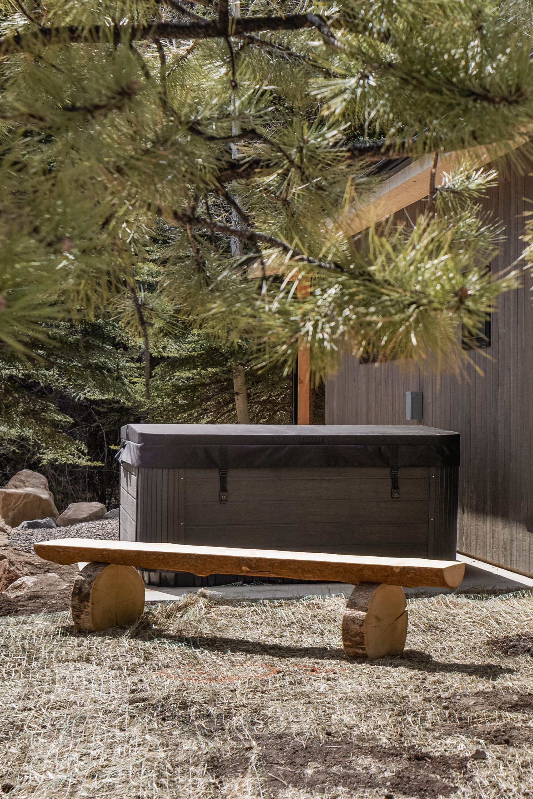 A wooden bench sits on dry grass in front of a dark hot tub with a cover, next to a wooden cabin, surrounded by pine trees in an outdoor, forested setting.
