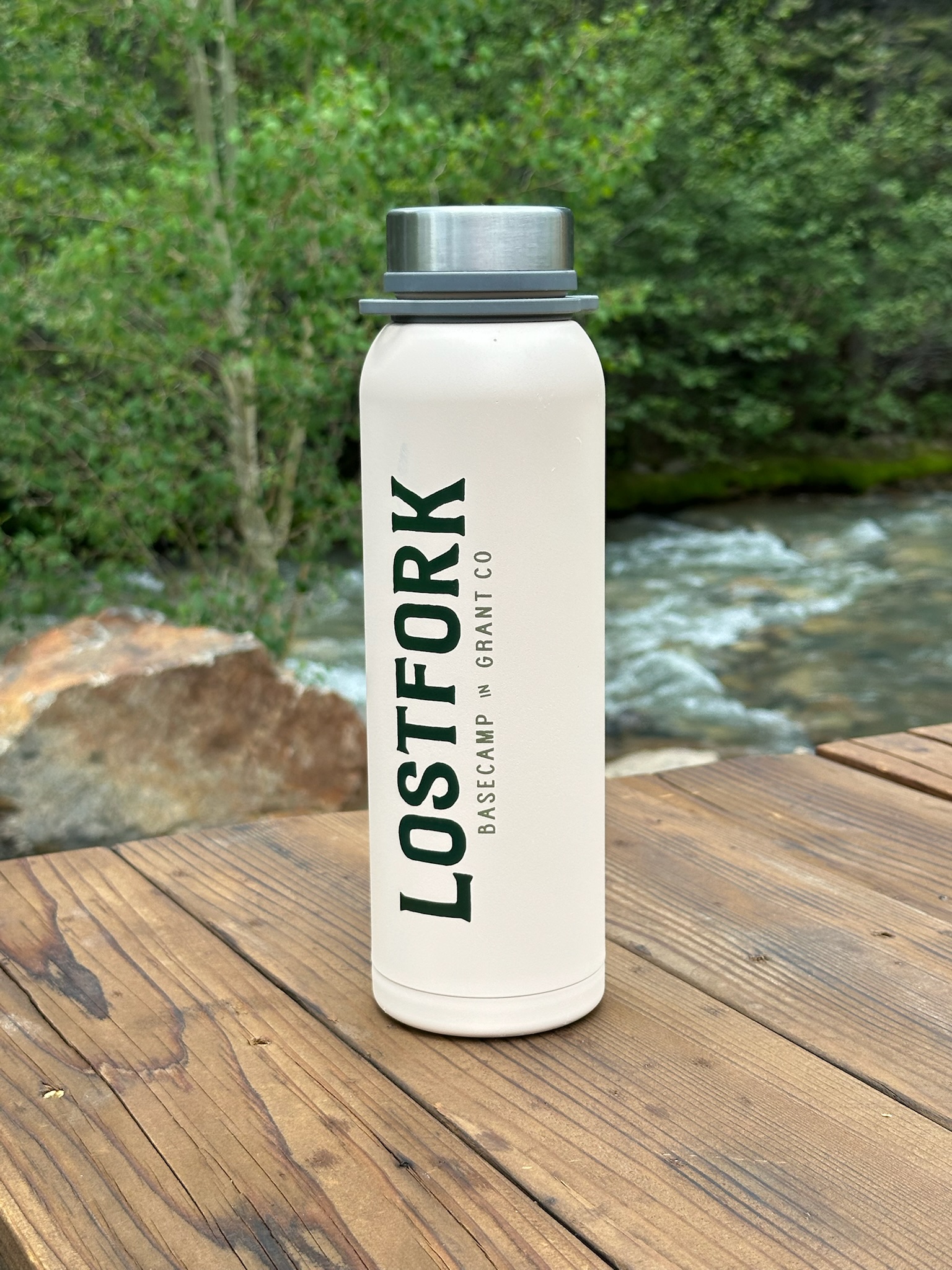 A white water bottle with the text "LOSTFORK BASECAMP BY GRANT CO" stands on a wooden surface outdoors, with a river, rocks, and green trees in the background.