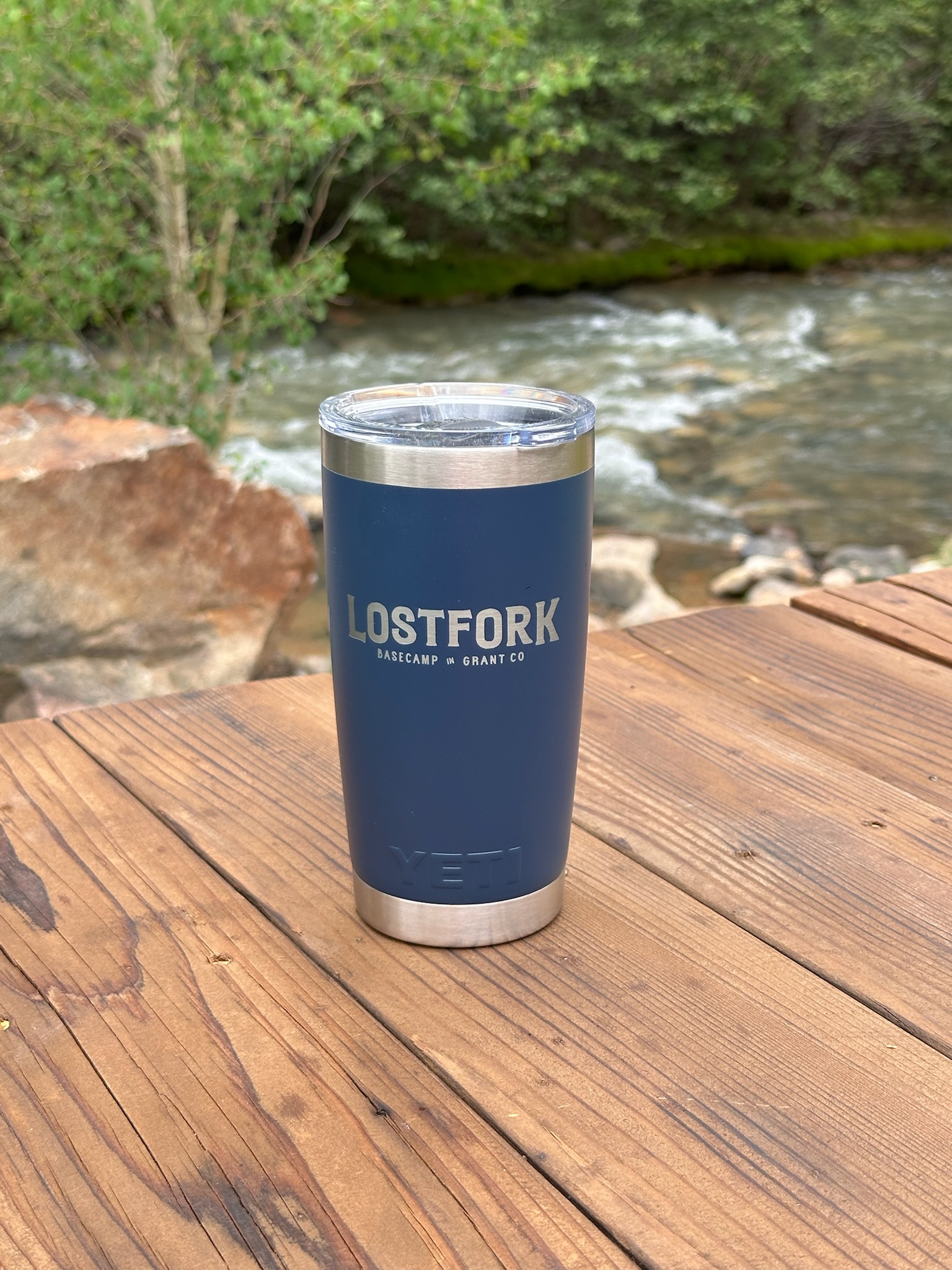 A blue stainless steel tumbler with “LOSTFORK BASECAMP + GRANT CO” written on it sits on a wooden table outdoors near a flowing river and trees in the background.