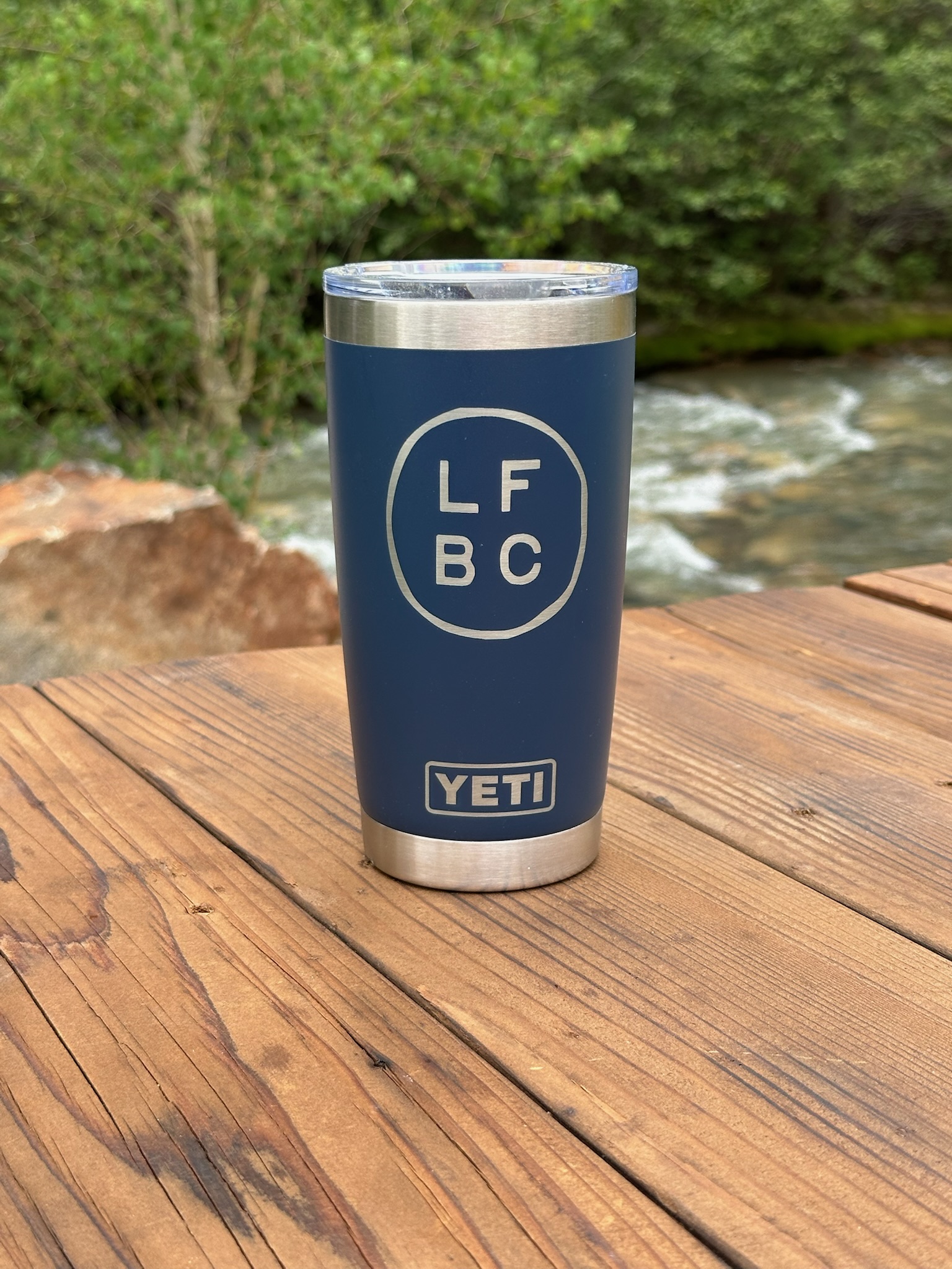 A blue YETI tumbler with “LFBC” in a white circle sits on a wooden table outdoors, with greenery and a flowing river in the background.